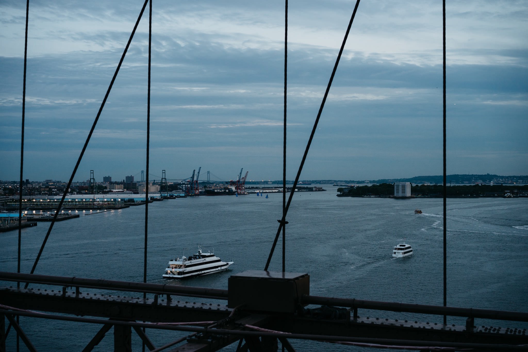 View of the East River in New York City from a bridge, showing two boats and the city skyline at dusk.