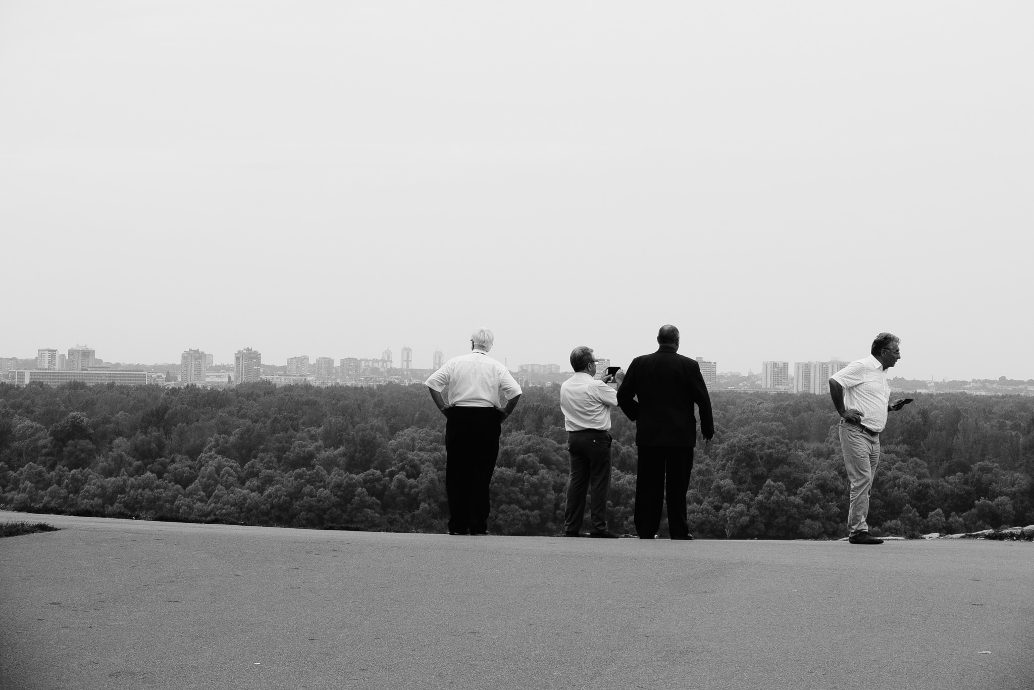 Four men stand overlooking a city skyline from a tree-lined hill.