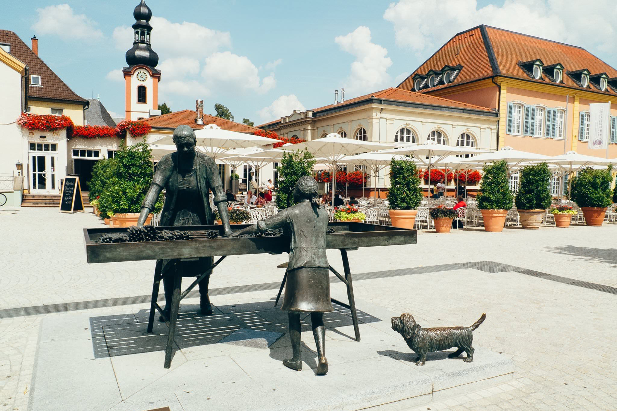 Bronze statue of two women and a dog selling asparagus in a town square.