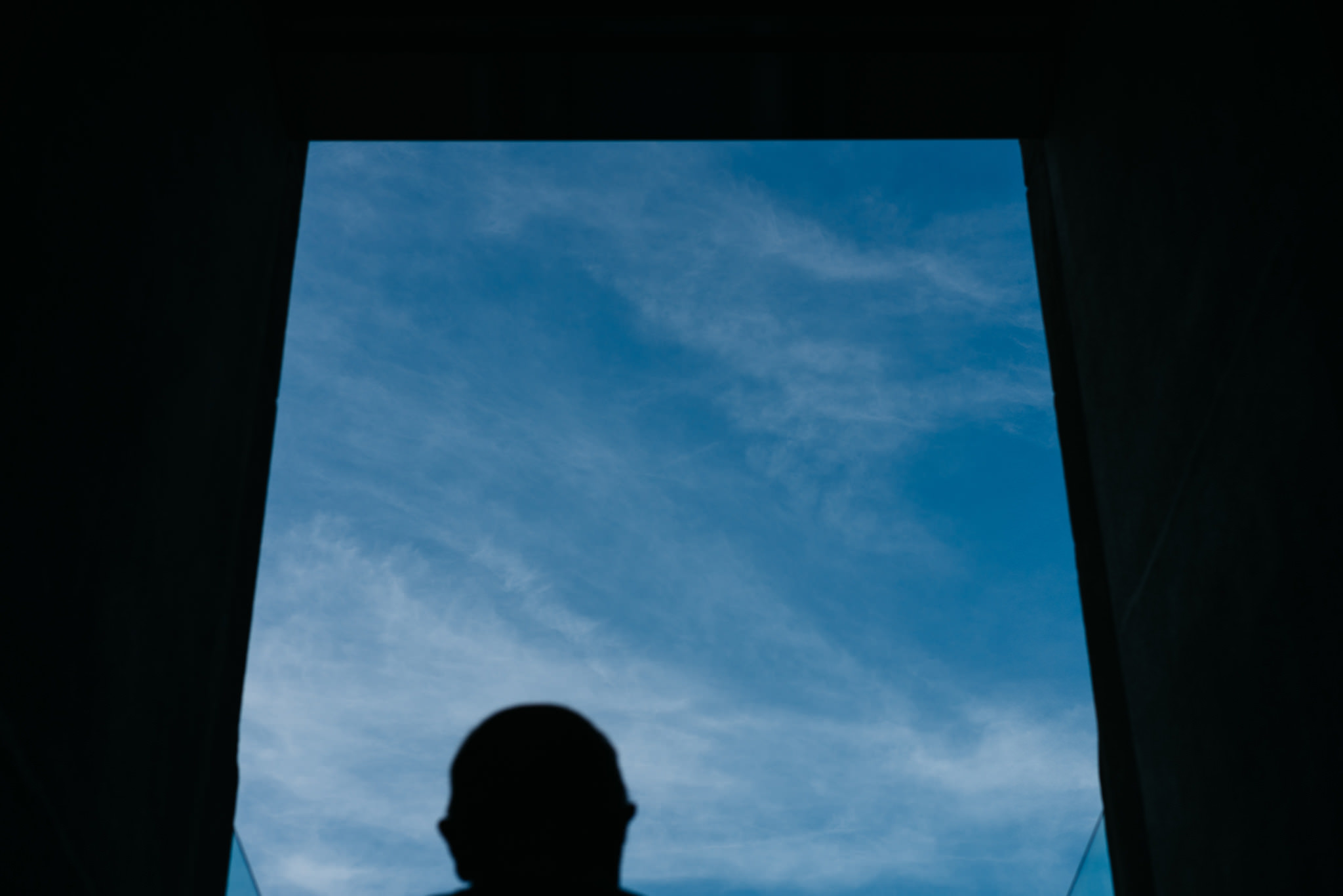 Silhouette of a person's head and shoulders viewed from below through a dark rectangular opening, against a bright blue sky with wispy clouds.