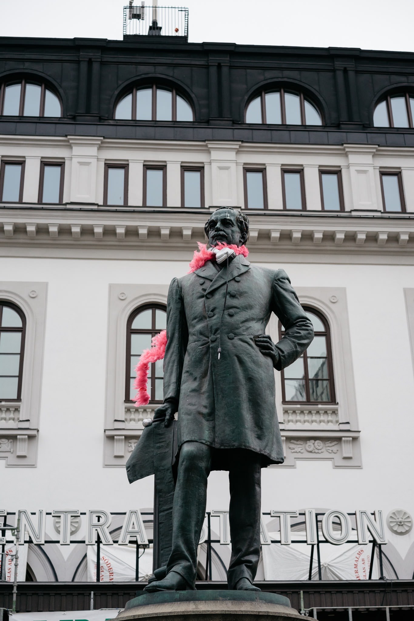 Statue of a man in a long coat adorned with pink boas, standing in front of a building.