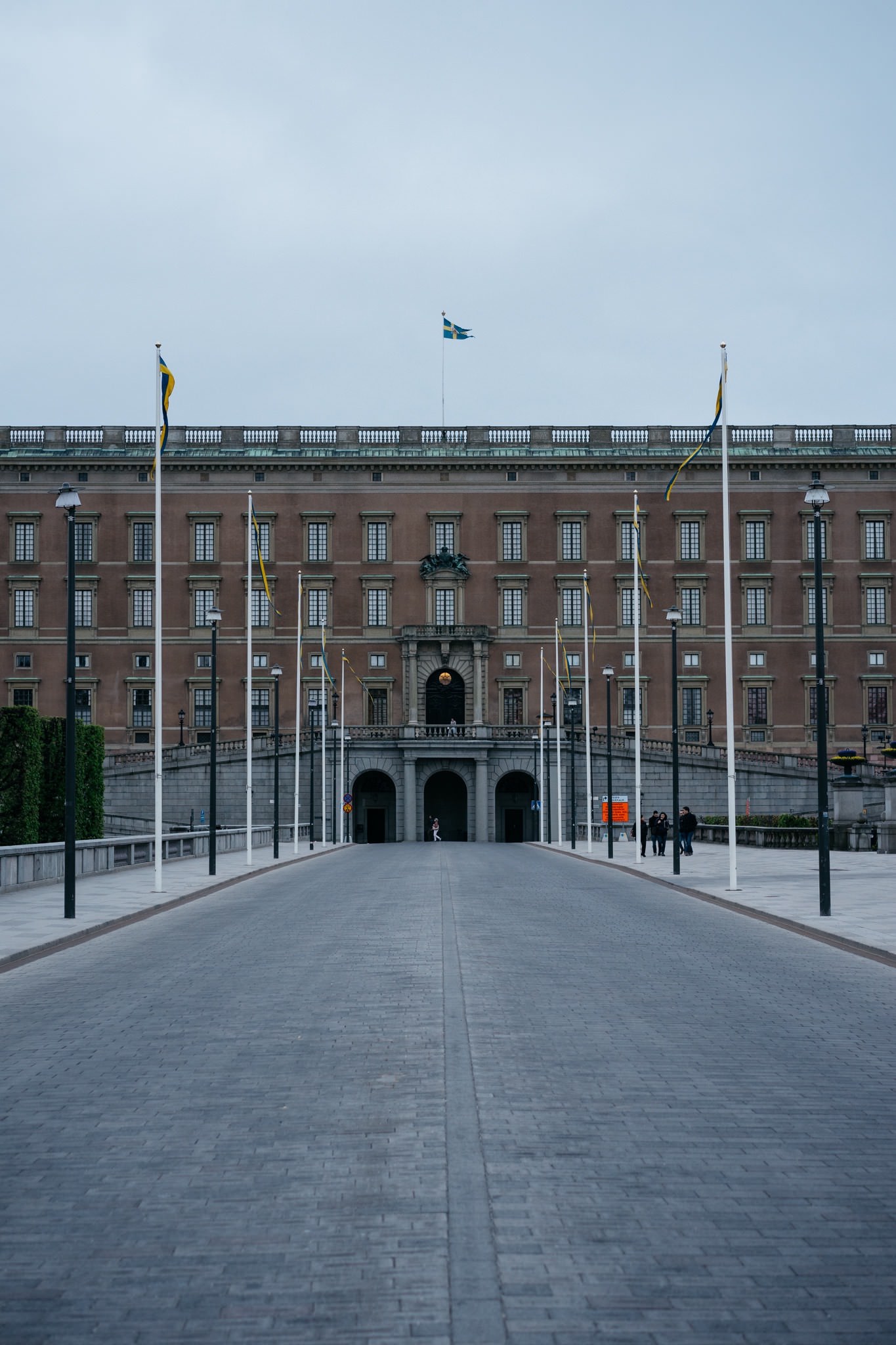 Stockholm Palace with Swedish flags, seen from a long perspective down a paved pathway.