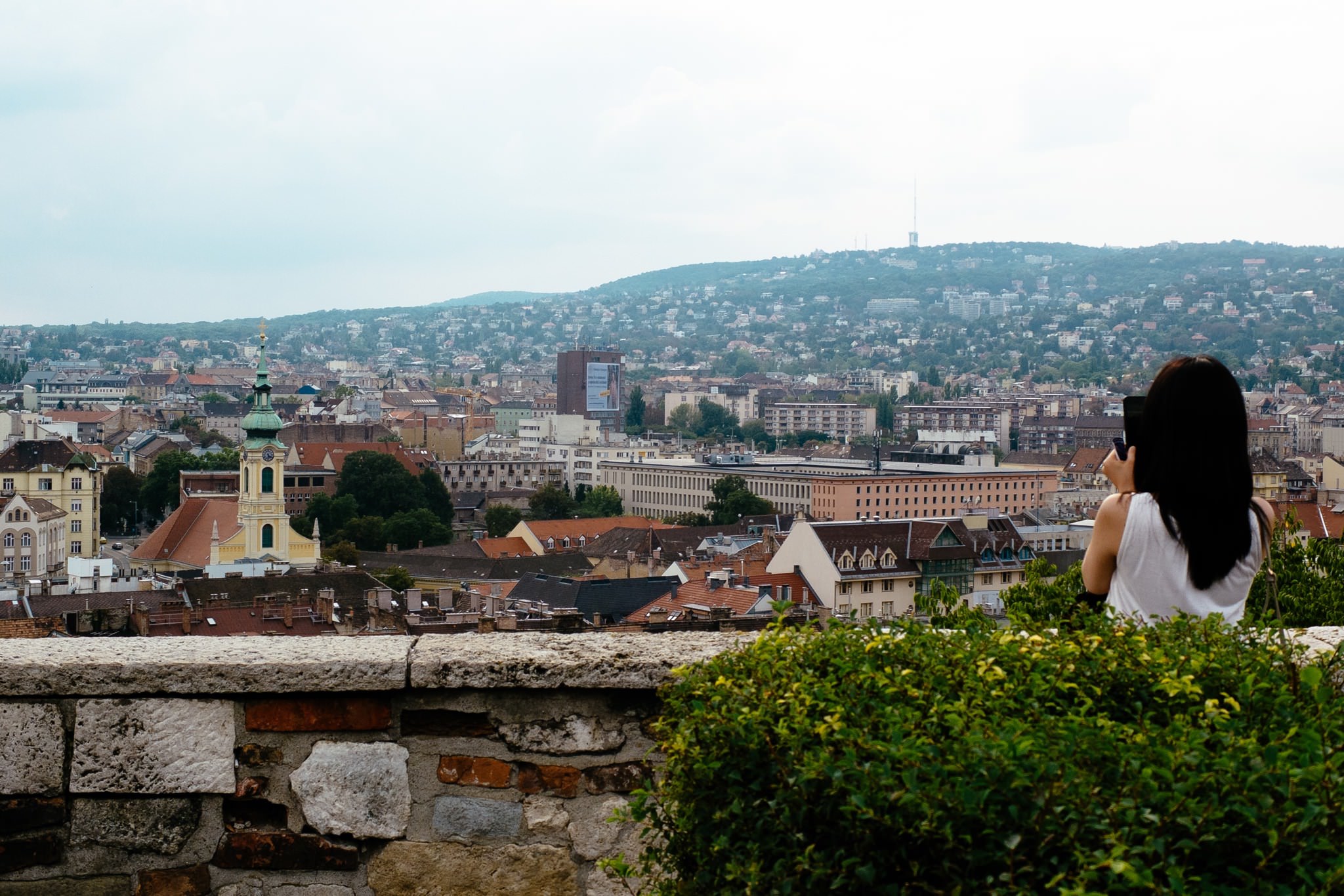 Woman taking a picture of Budapest cityscape from behind a stone wall.