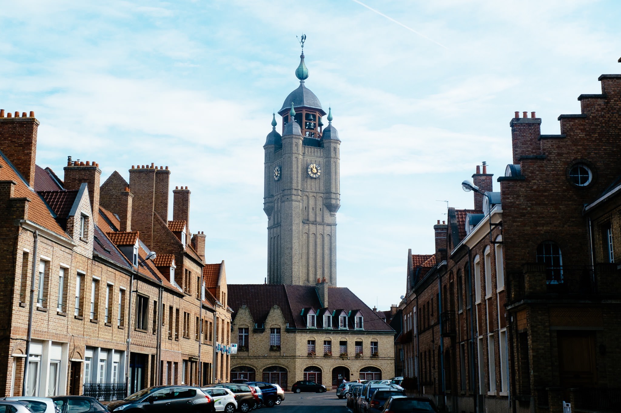 Tall clock tower in Bergues, France, seen from a street lined with brick buildings.