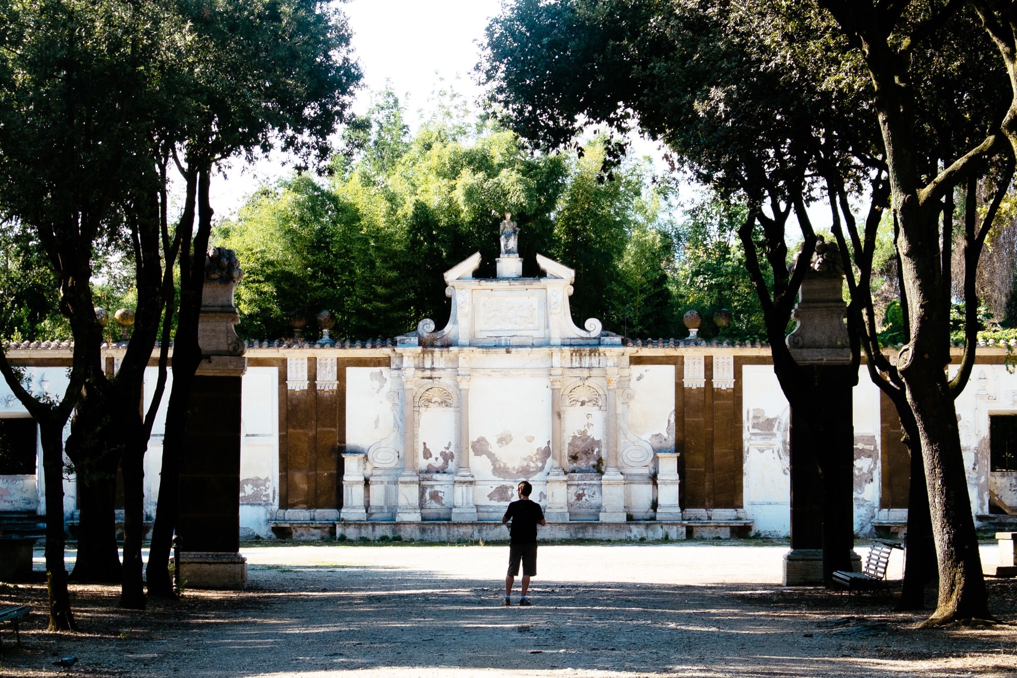 Person standing before a weathered, ornate stone wall in a park.