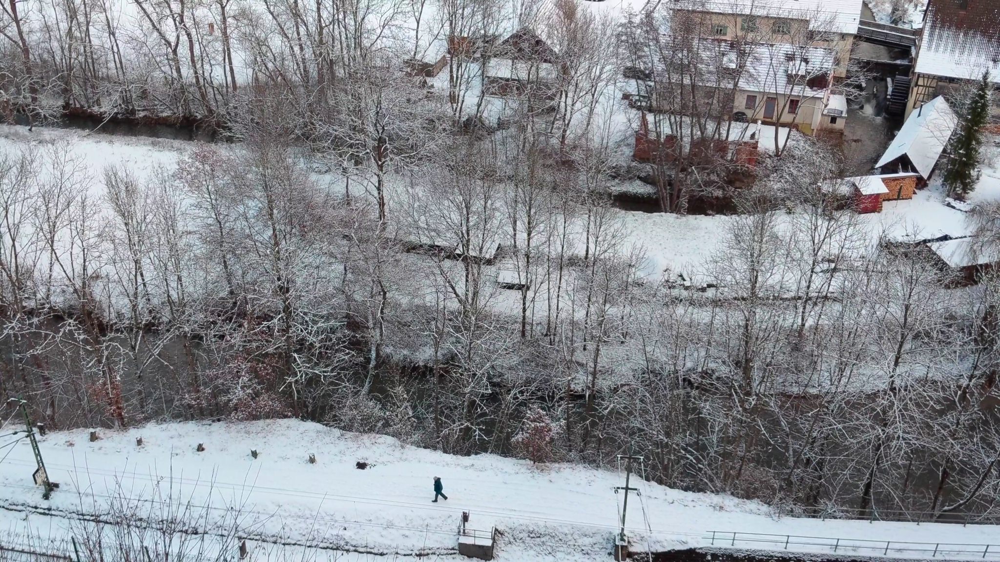 Aerial view of a snow-covered village with a person walking along a snowy path by a river.