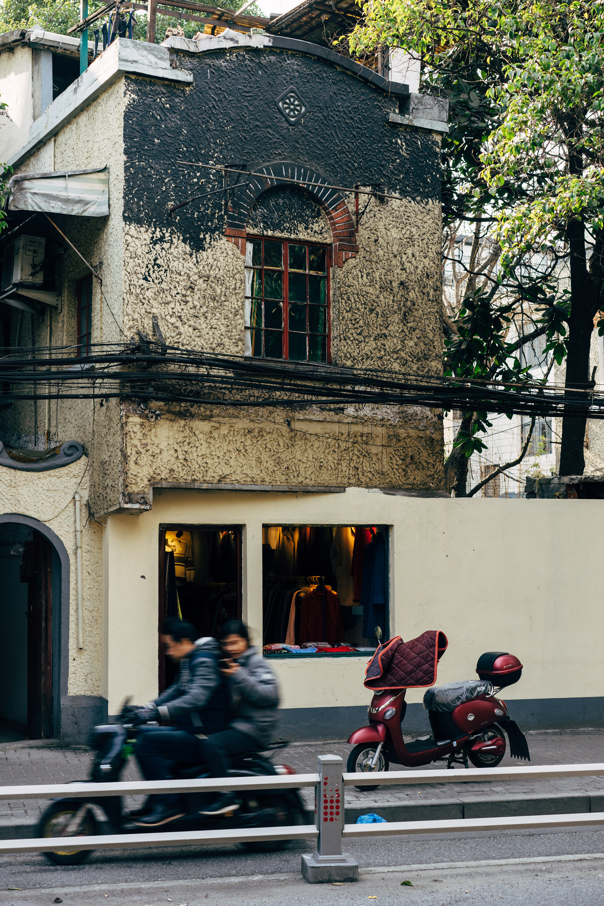 Shanghai street scene: a weathered building with a shop displaying clothes, a parked red scooter, and a person riding a scooter past.