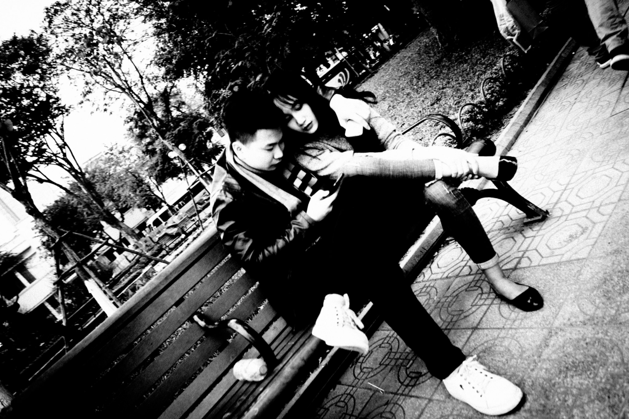 Black and white photo of a young couple sitting on a bench, looking at a phone.