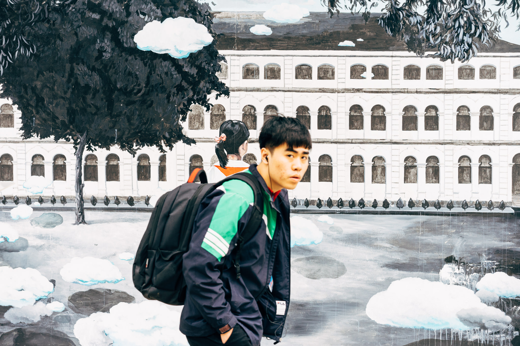 Young man with backpack standing in front of a mural depicting a building and clouds.