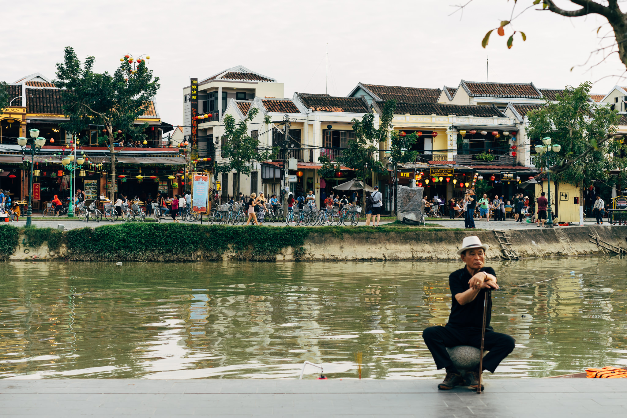 A man sits on a stone by a river, in front of shops and people in Hoi An.