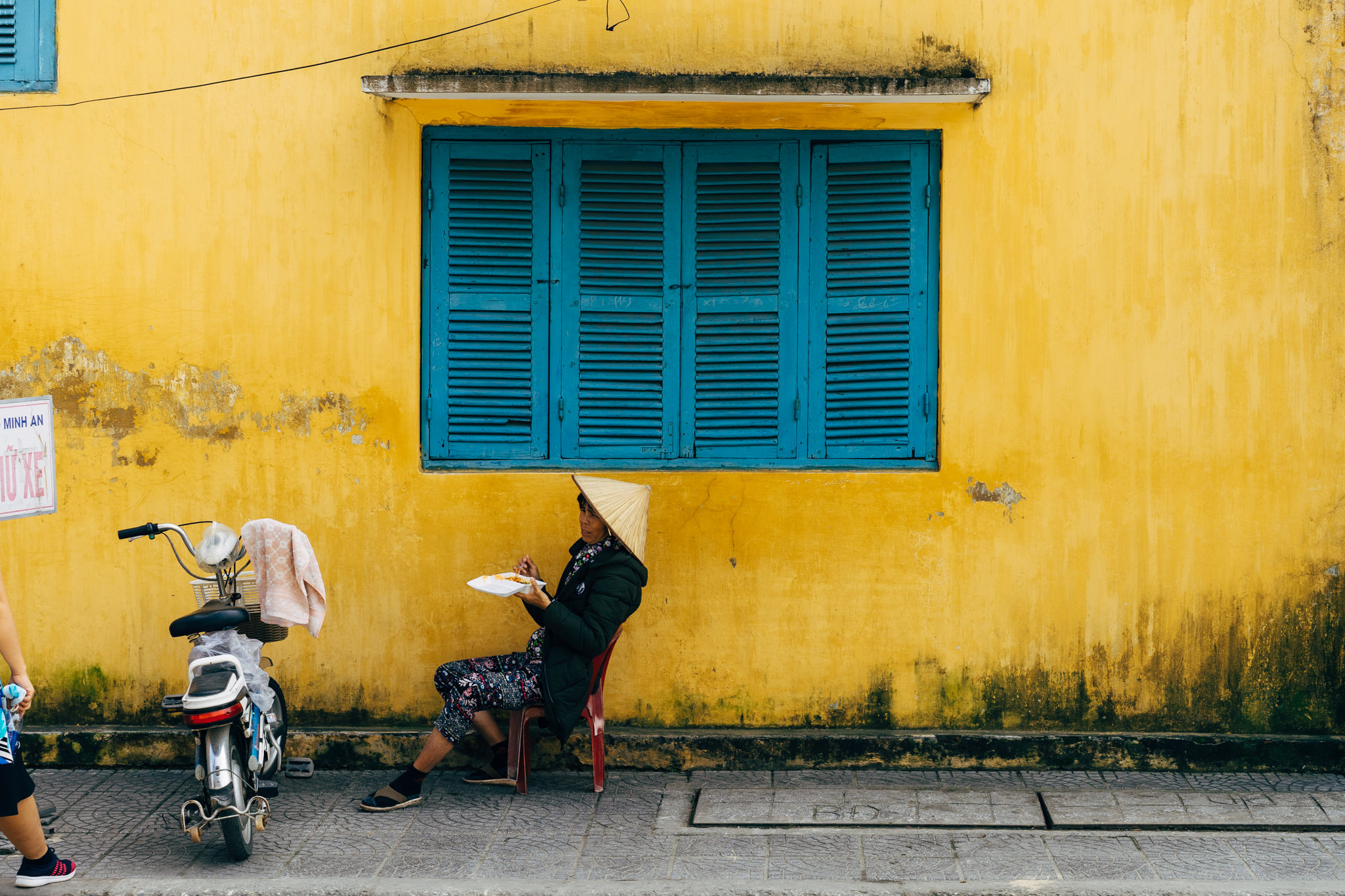 Woman in conical hat eating outside a yellow building with blue shutters.