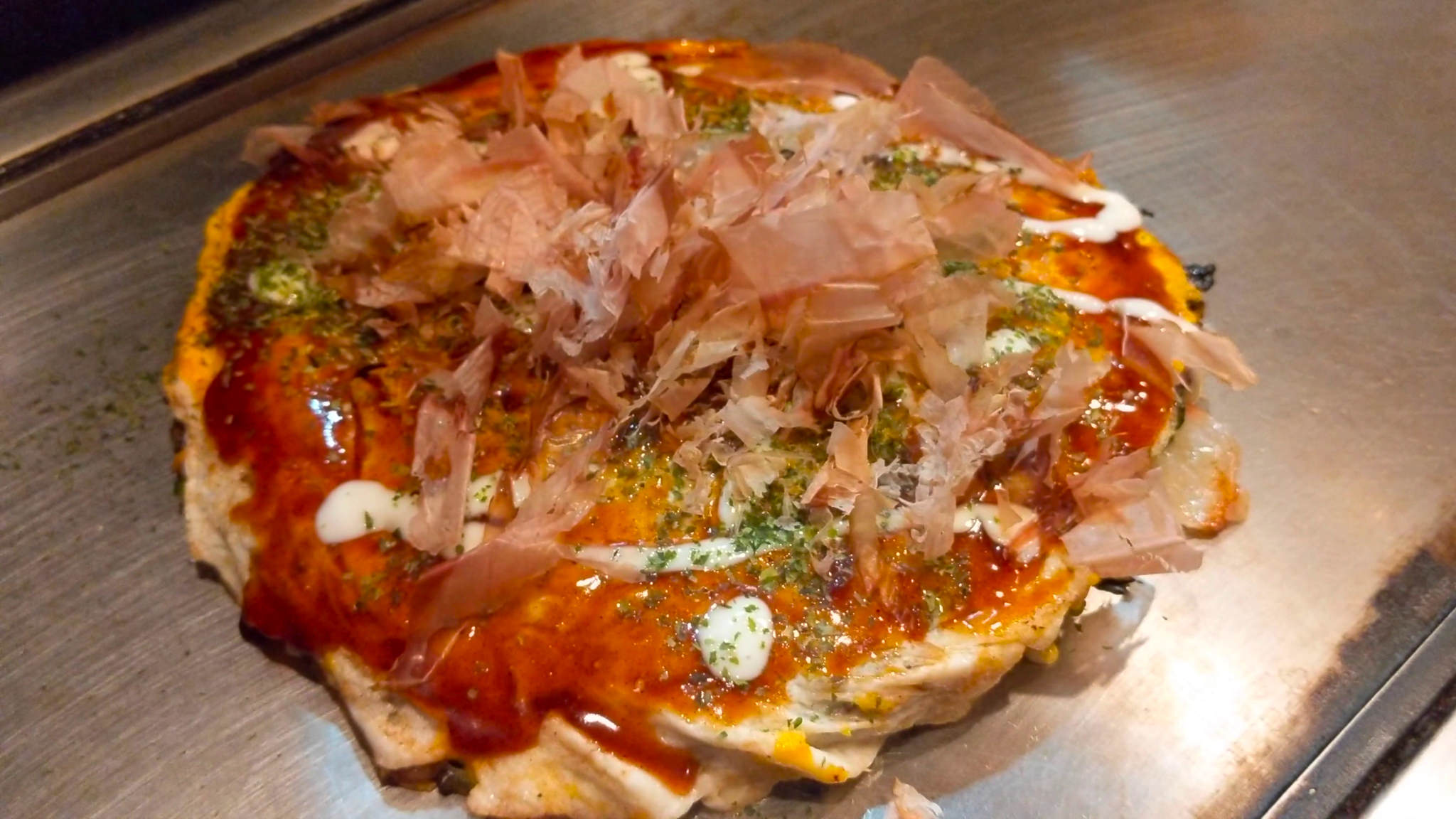 Okonomiyaki topped with bonito flakes and mayonnaise on a griddle.