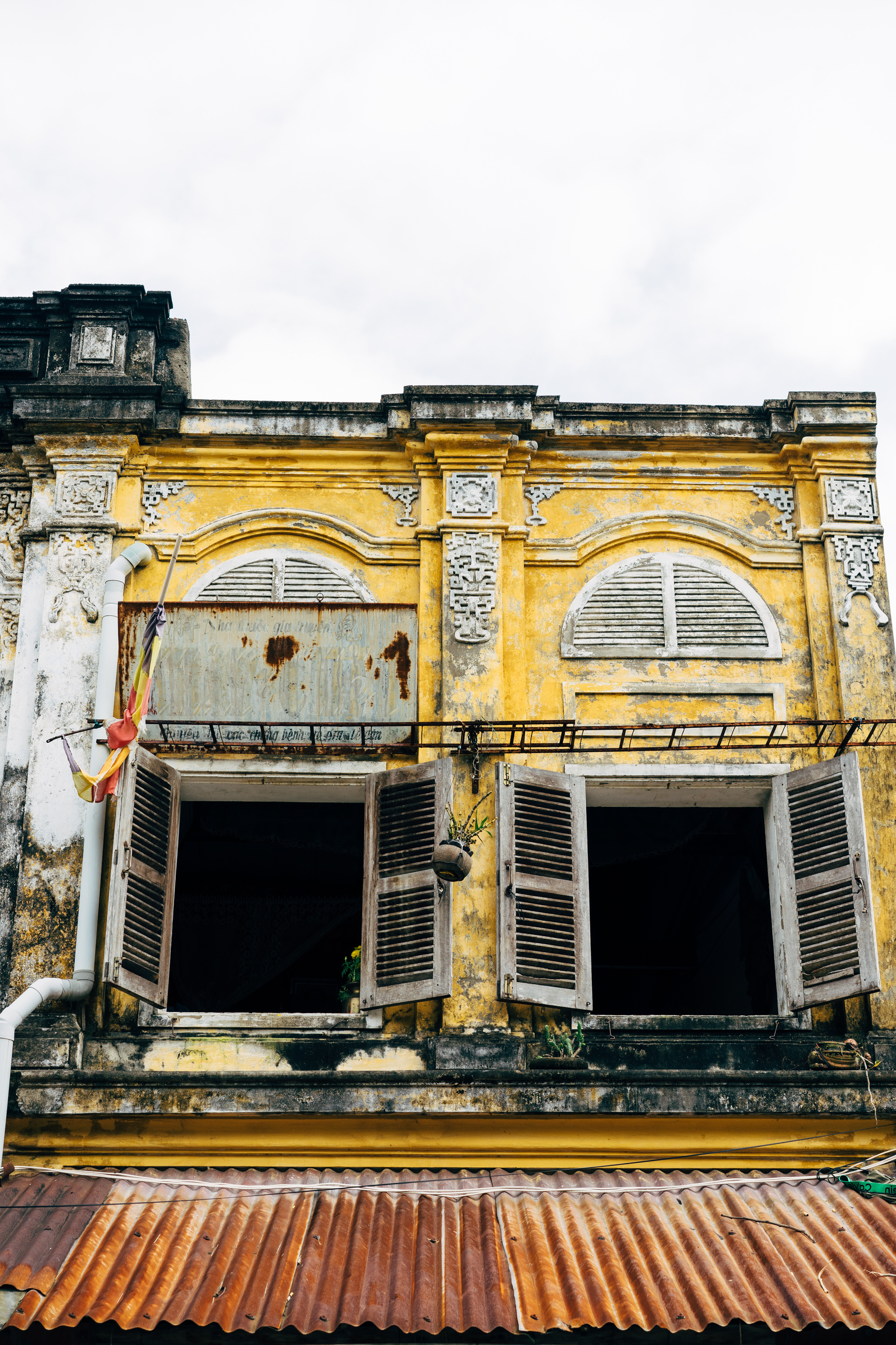 Old Hoi An building facade with yellow walls, open wooden shutters, and rusty corrugated metal roof.