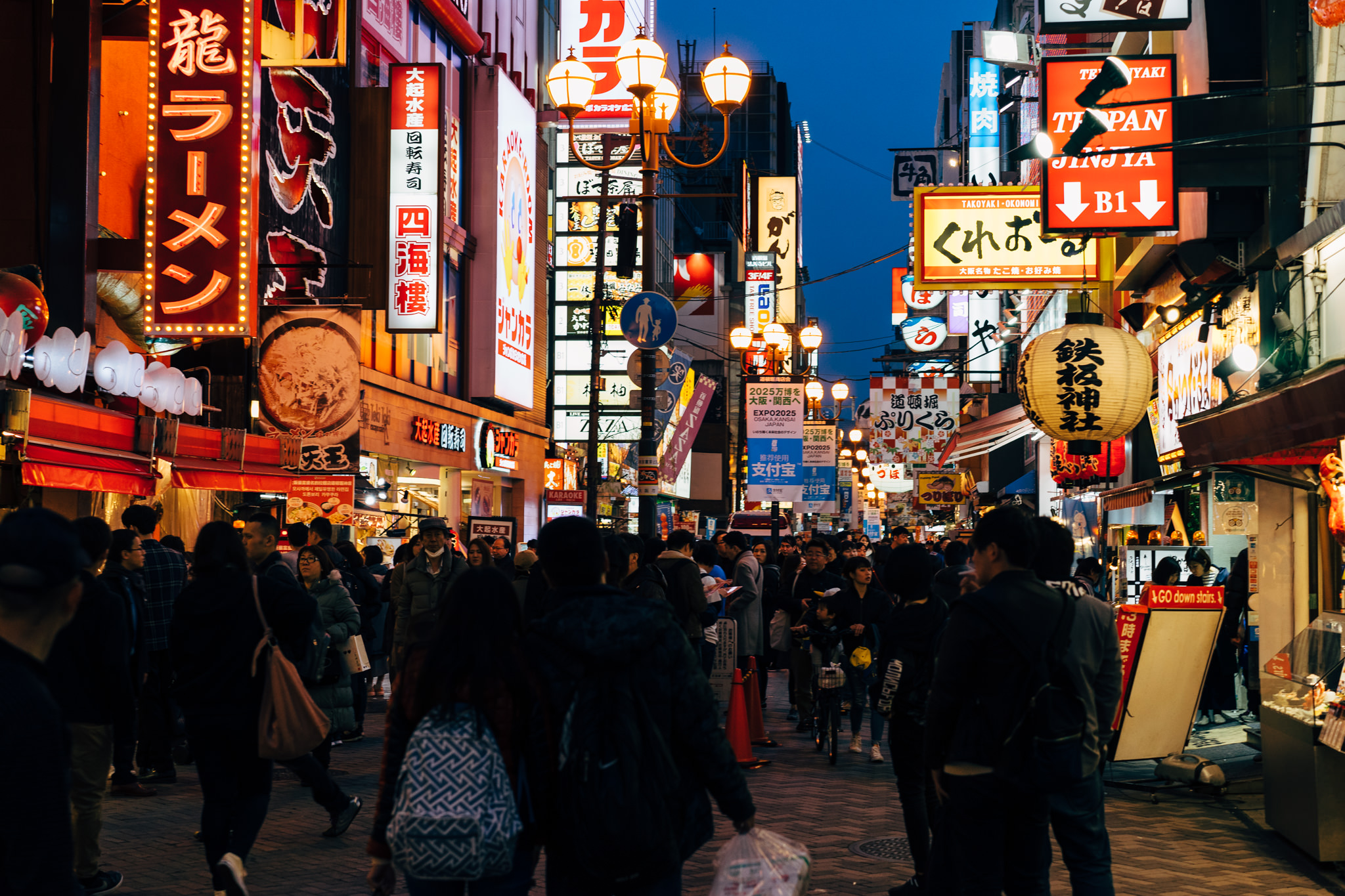 Night view of a crowded street in Osaka, Japan, with illuminated signs in Japanese and English.