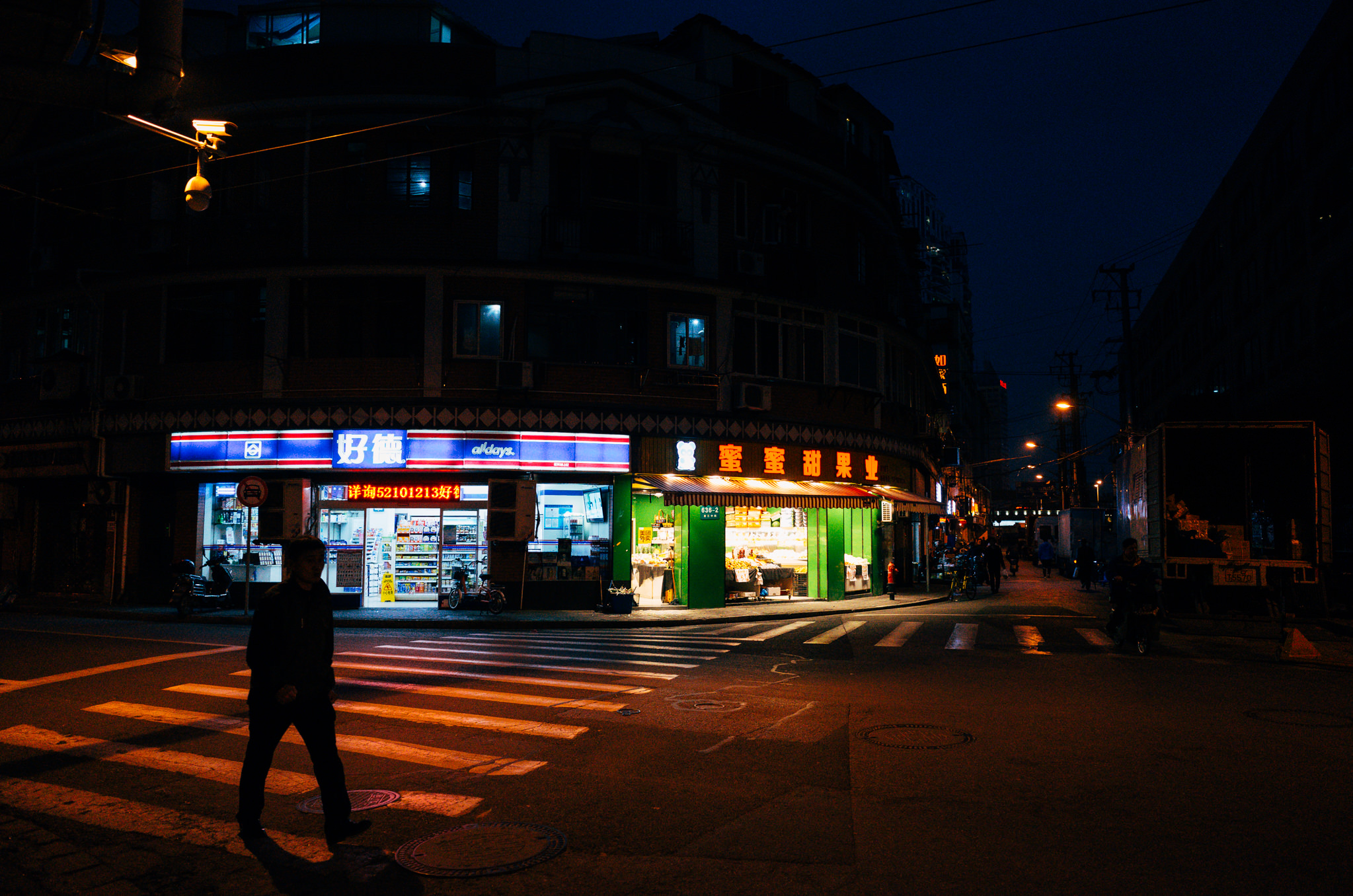 Nighttime street scene in Shanghai, China, showing a convenience store and fruit stand with pedestrians crossing the street.