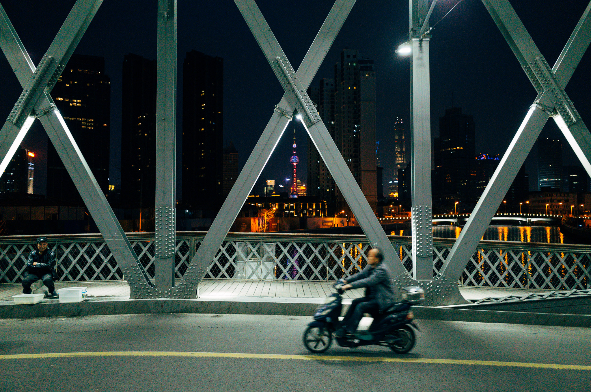 Person on motor scooter on Shanghai bridge at night, city skyline in background.