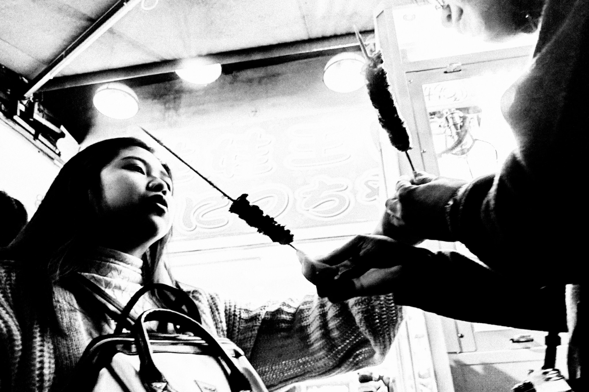 Black and white photo of a young woman and a person sharing street food on a skewer.