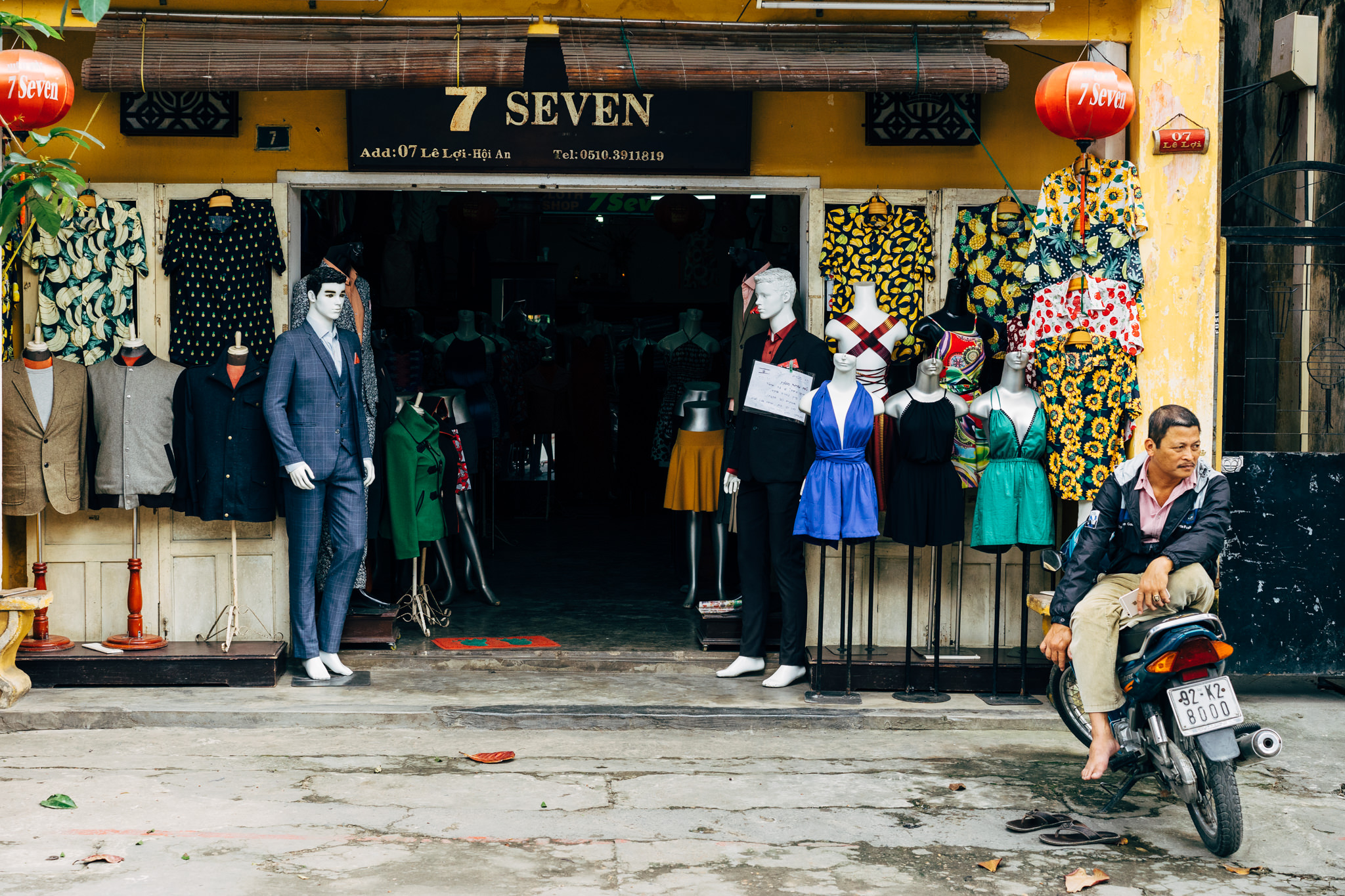 A man sits on a motorbike outside a Hoi An clothing shop, '7 Seven,' displaying men's and women's clothing.