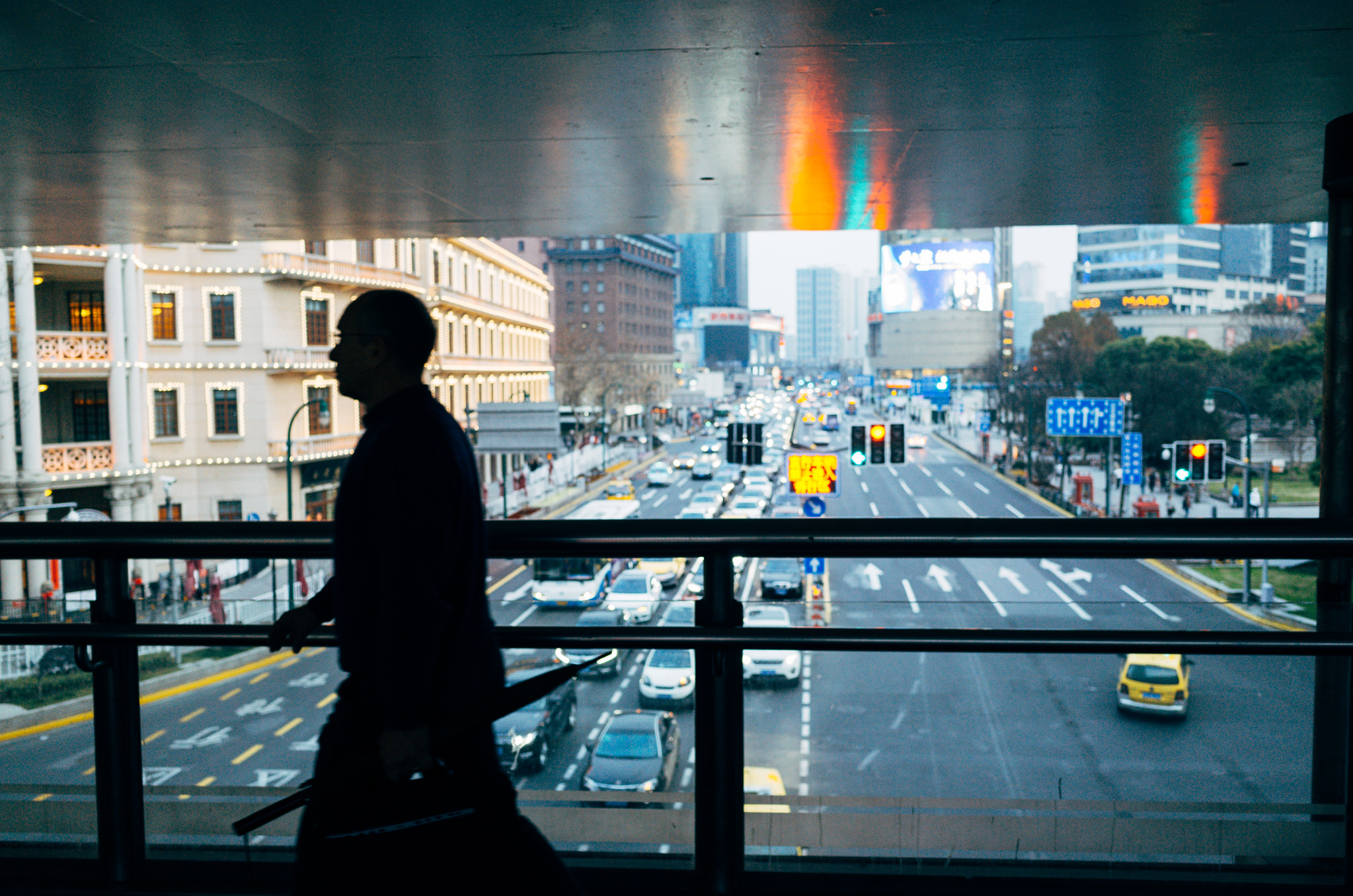 Silhouette of a man walking on a pedestrian overpass overlooking a busy Shanghai street at night.