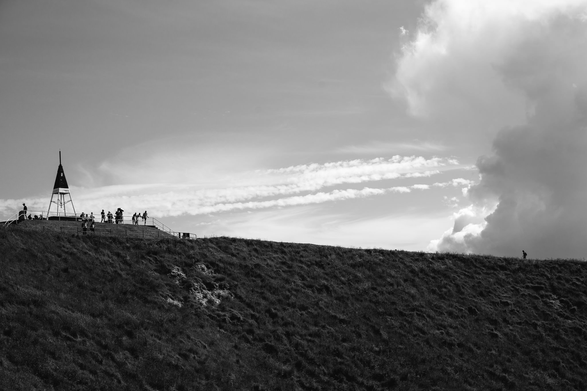 Black and white photo of people standing on a hilltop viewing platform near a small tower, under a partly cloudy sky.