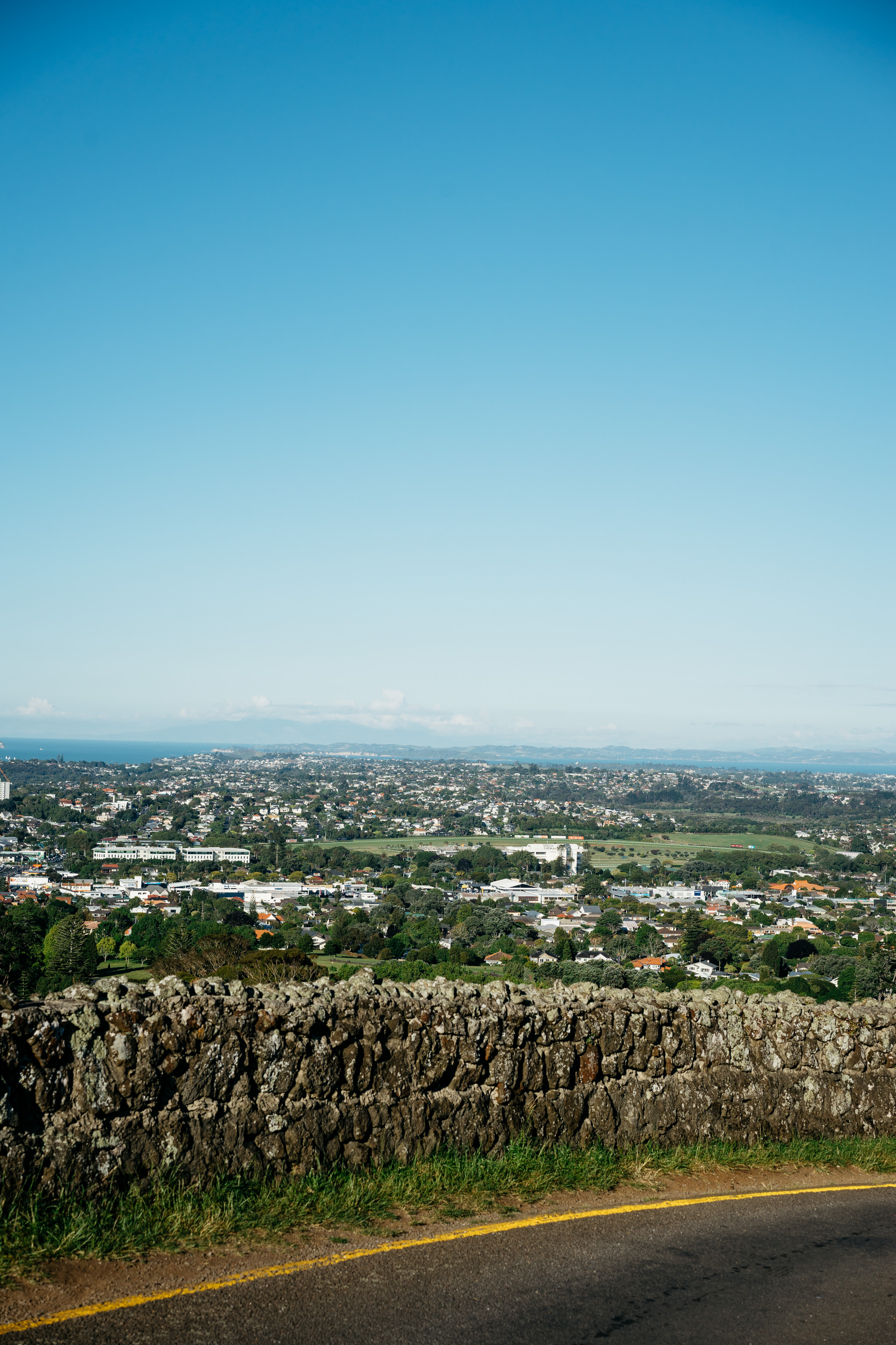 Elevated view of Auckland, New Zealand, seen from behind a low stone wall next to a road.