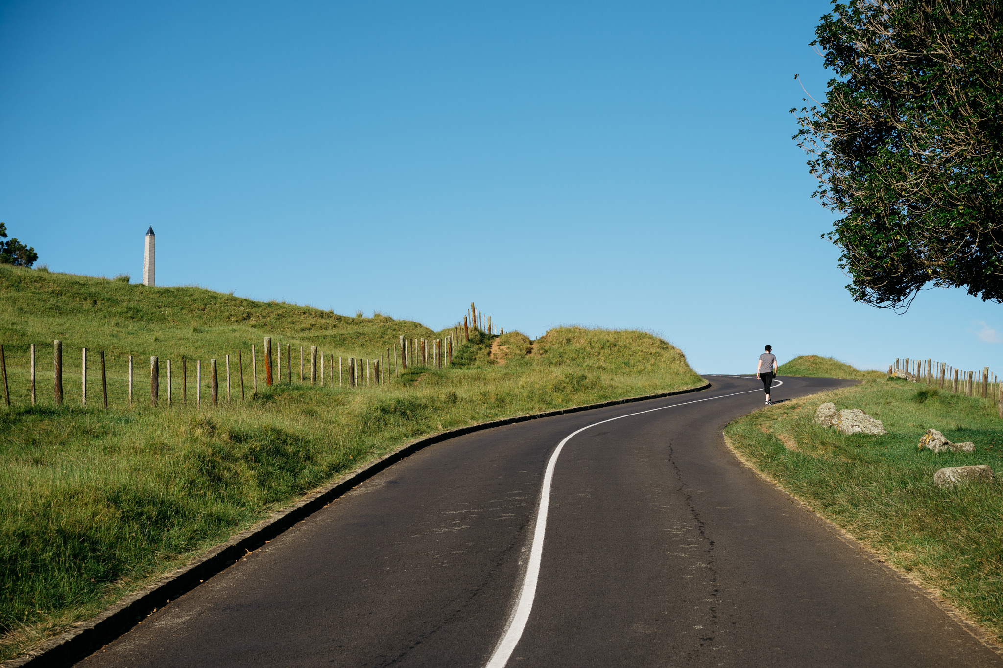 Person walking away on a winding road with grassy hills and a monument in the background.