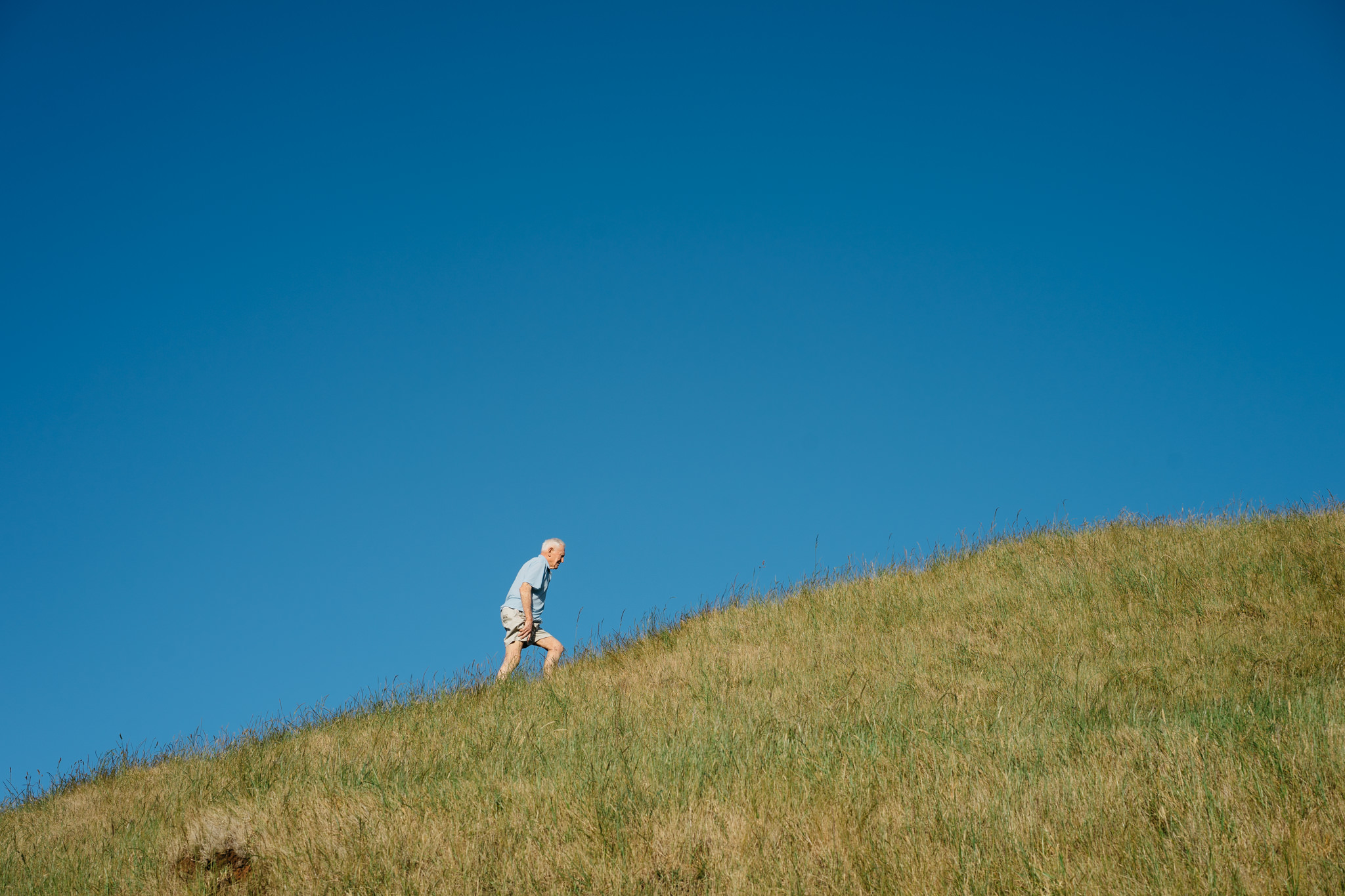 Elderly man walking uphill in grassy field under a clear blue sky.