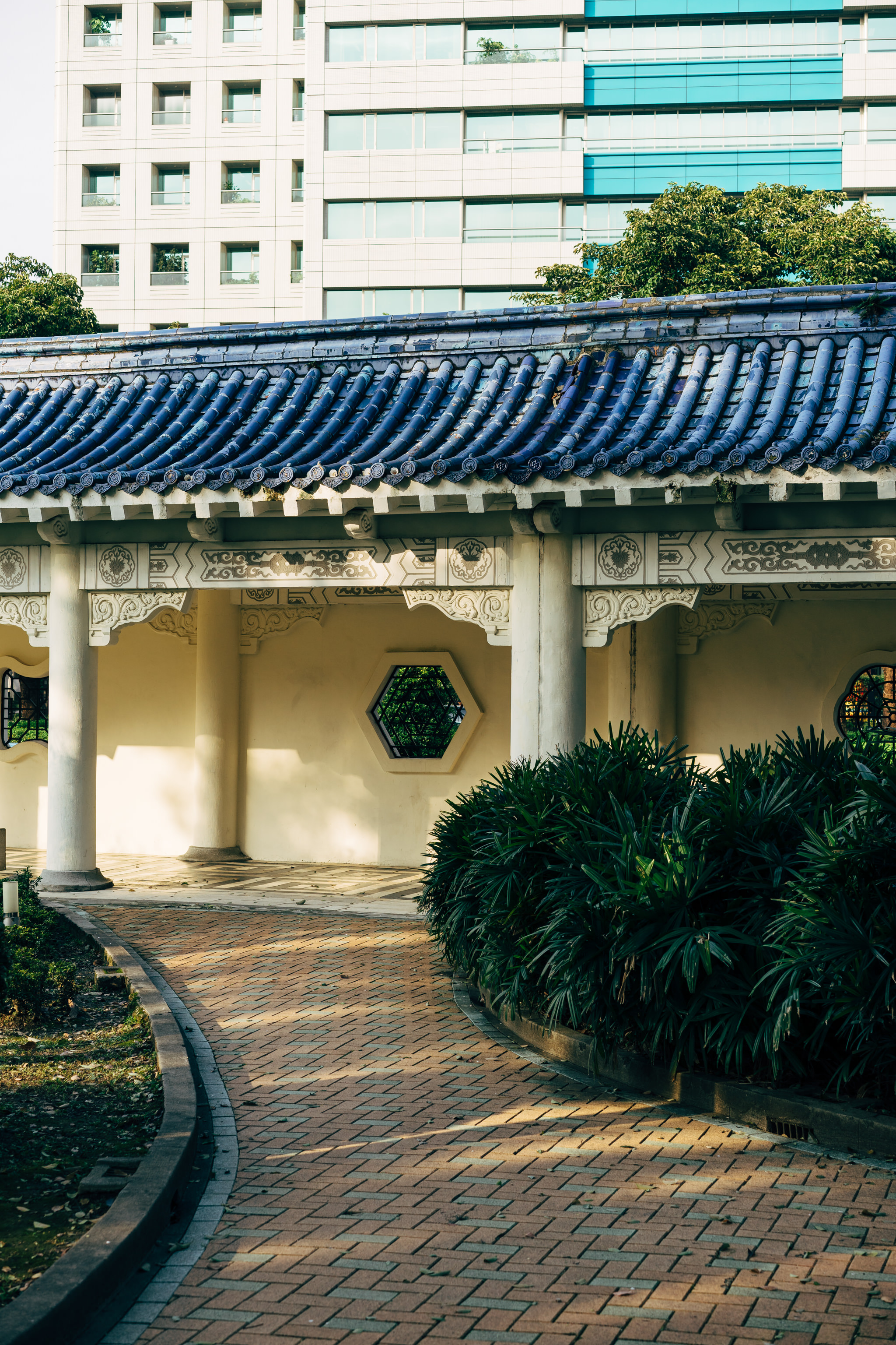 Brick pathway leading to a Chinese-style pavilion with a blue tiled roof, next to a modern building.