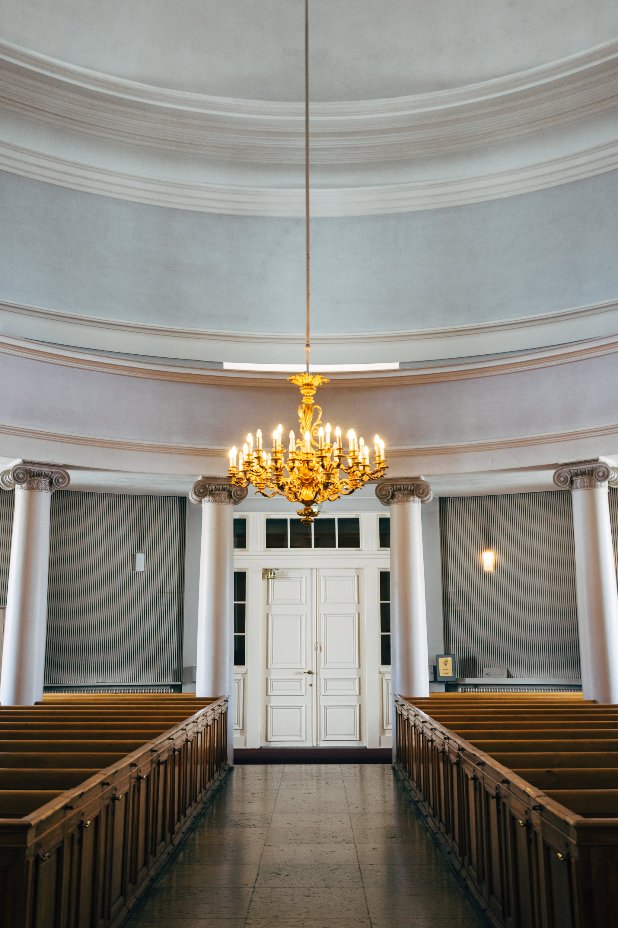 Interior of Helsinki Cathedral, showing a large golden chandelier and rows of wooden pews leading to a white door.