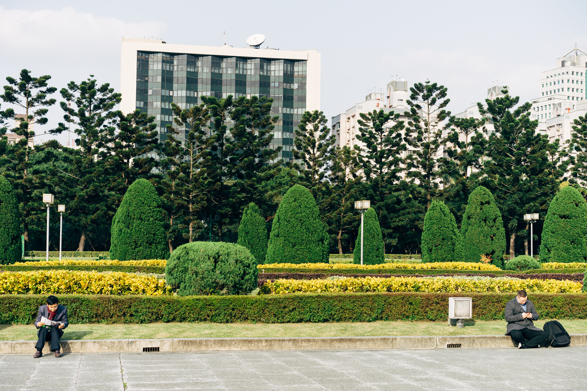 Two men sitting in Liberty Square, Taipei, amidst manicured hedges and tall buildings.