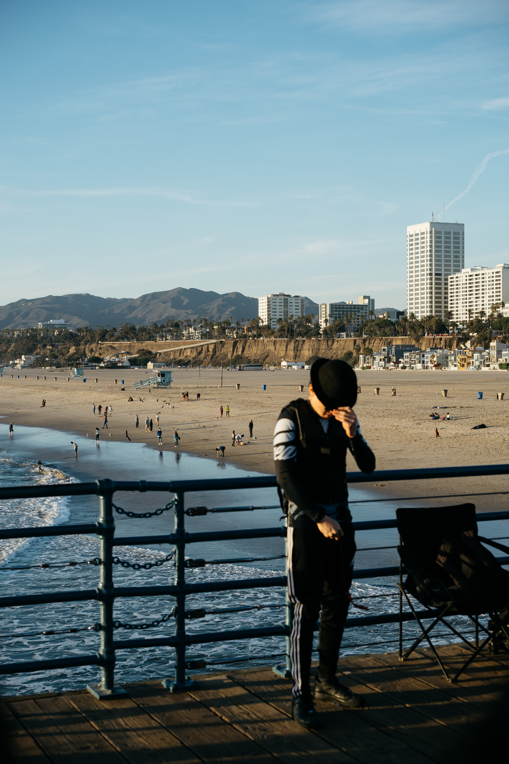 Person in hat standing on a pier overlooking a California beach and city skyline.