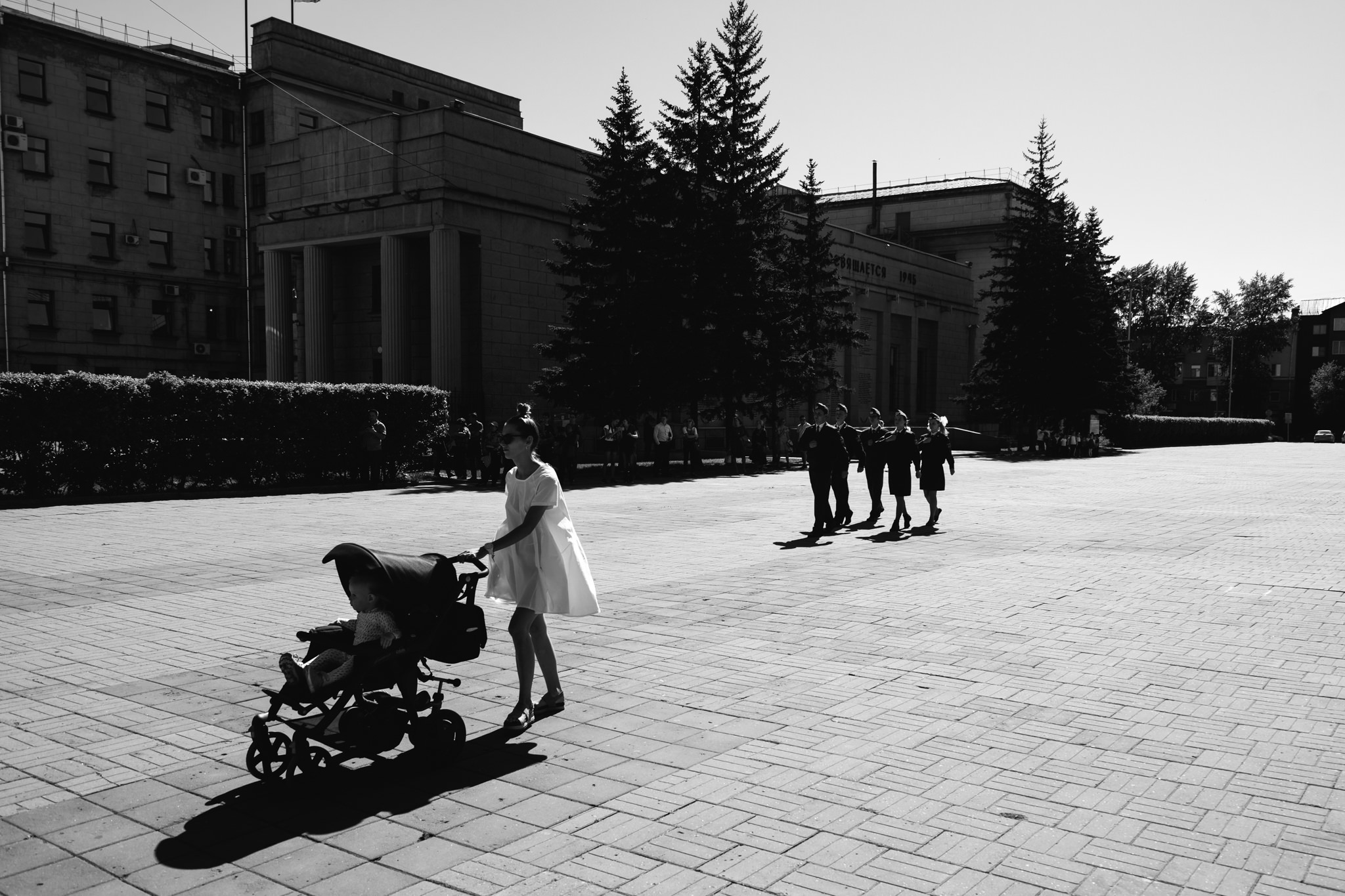 Black and white photo of a woman pushing a stroller past a group of people in uniform.