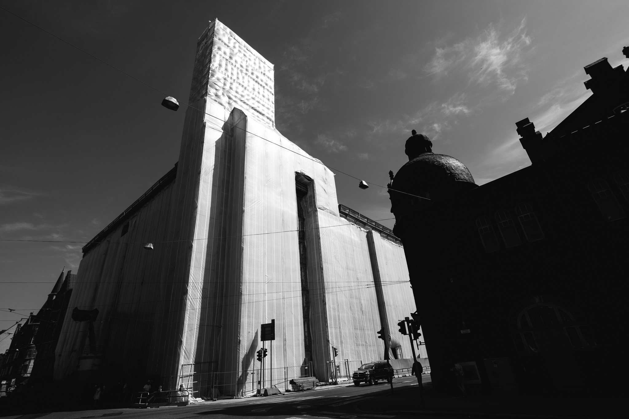 Black and white photo of a building wrapped in protective sheeting during construction, next to a dark building with a dome.