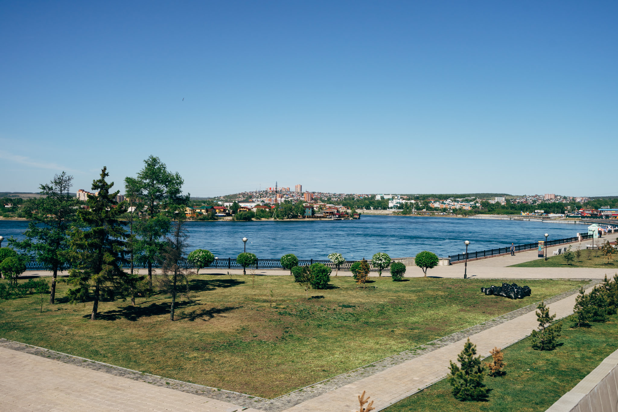 Irkutsk cityscape across the Angara River, viewed from a park with paved walkways.