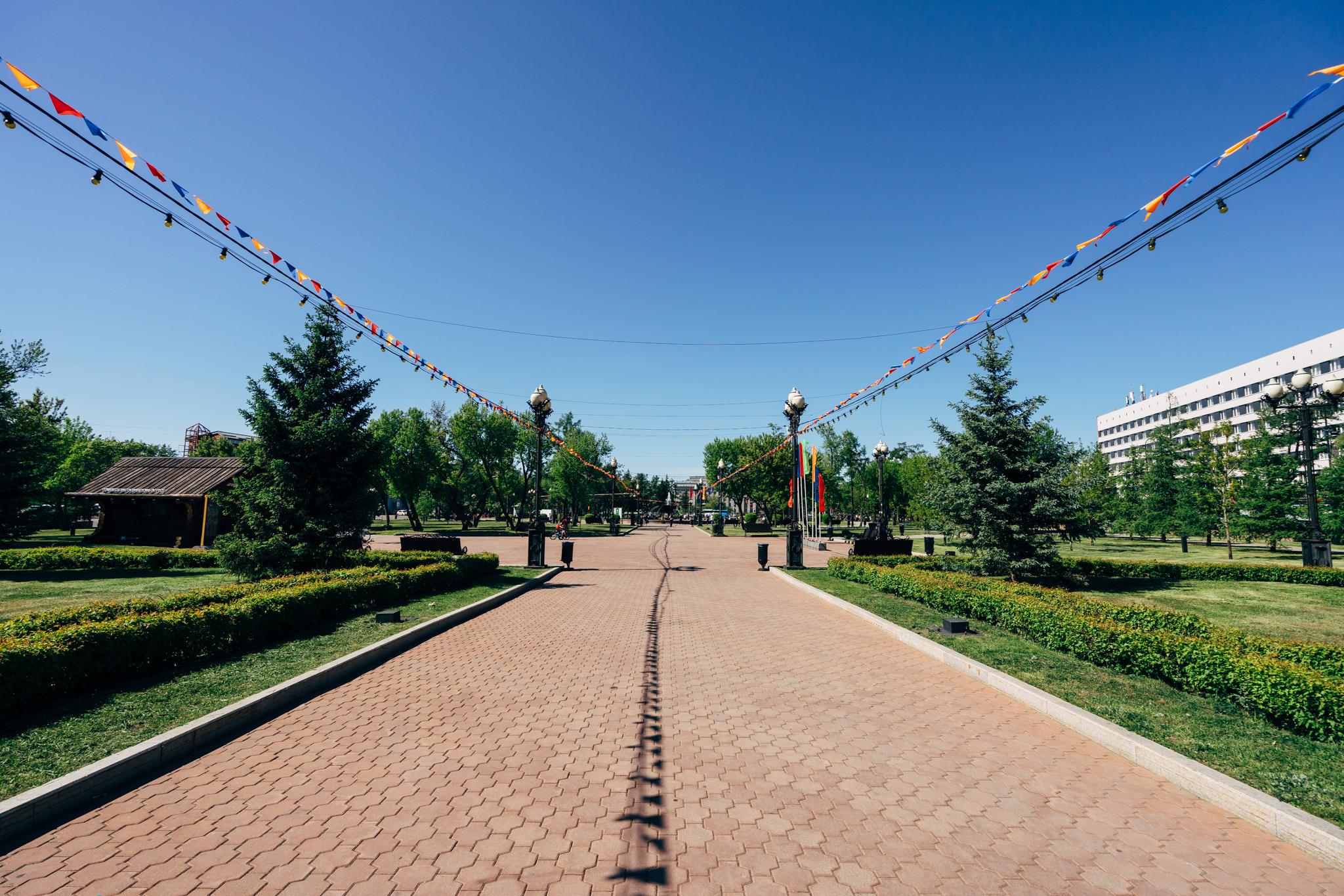 Park in Irkutsk, Russia, with a brick pathway lined with hedges and festive flags strung overhead.