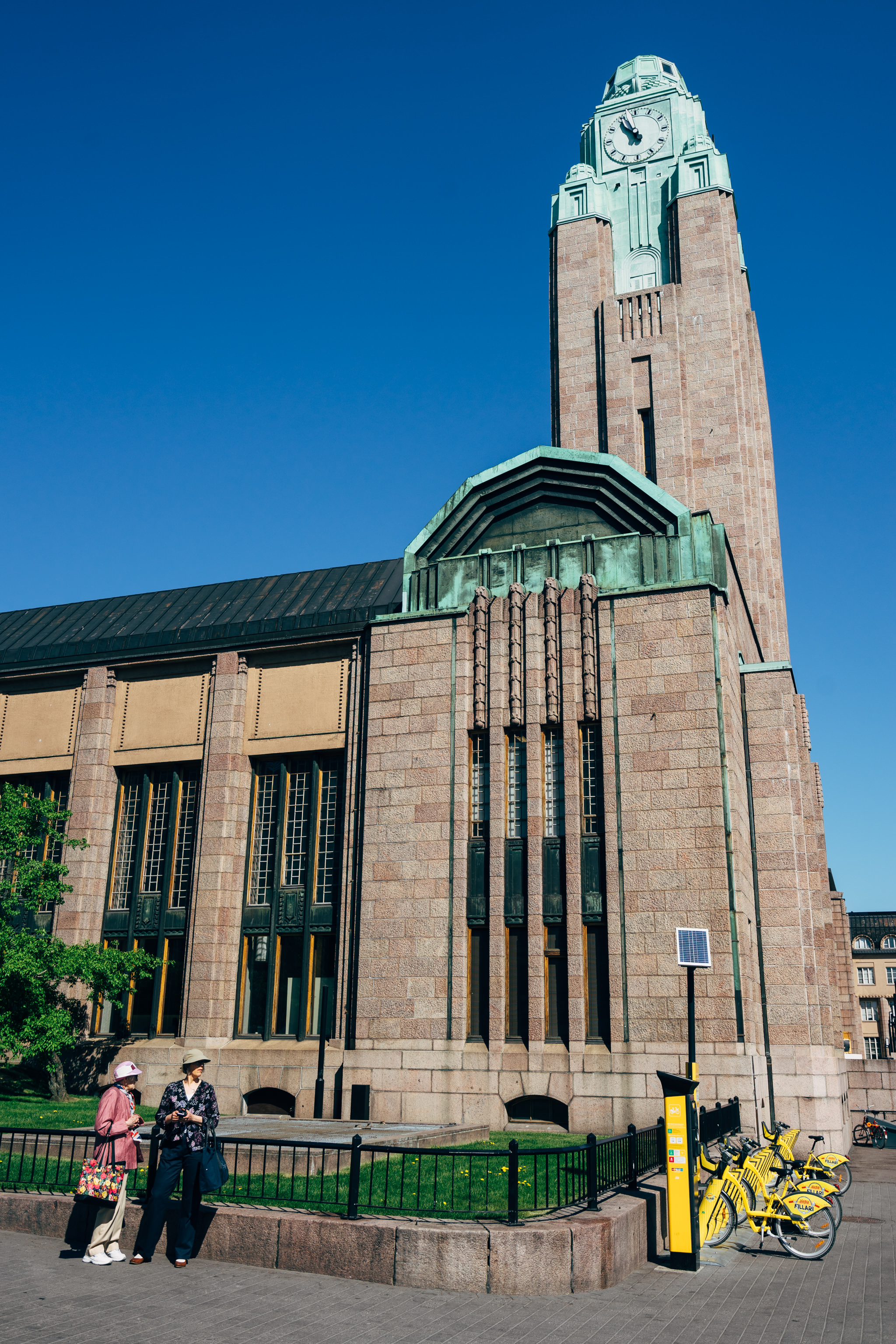 Helsinki Central Station with clock tower, two women standing nearby, and city bikes.