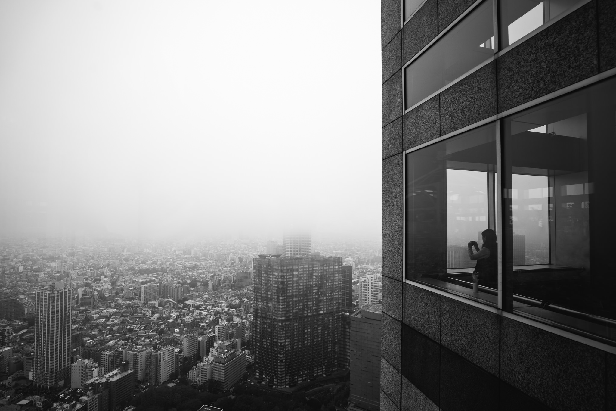 Black and white photo of a person taking a picture from a skyscraper observation deck overlooking a hazy Tokyo cityscape.