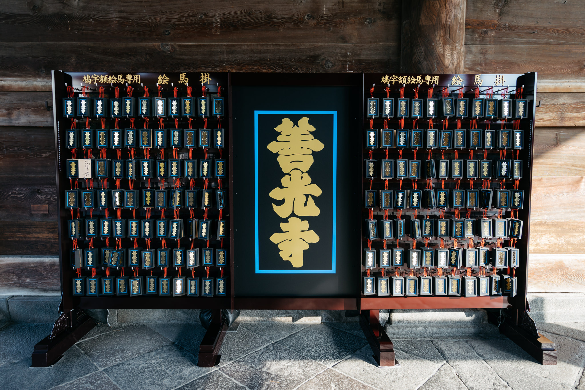 Wooden display with numerous small, dark-framed plaques containing gold lettering, flanking a larger central plaque with gold kanji characters.