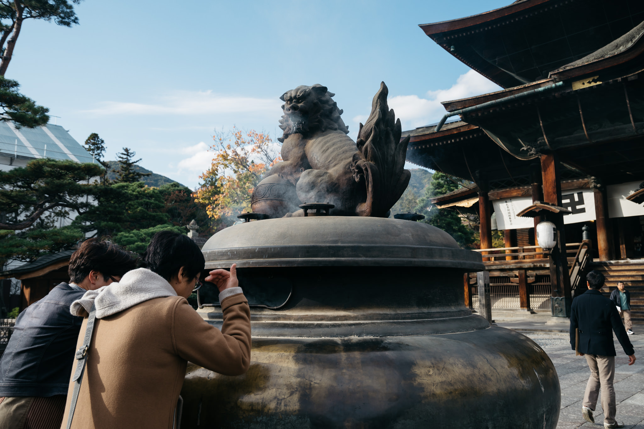Two people facing a large incense burner with a komainu statue at Zenko-ji Temple in Nagano, Japan.