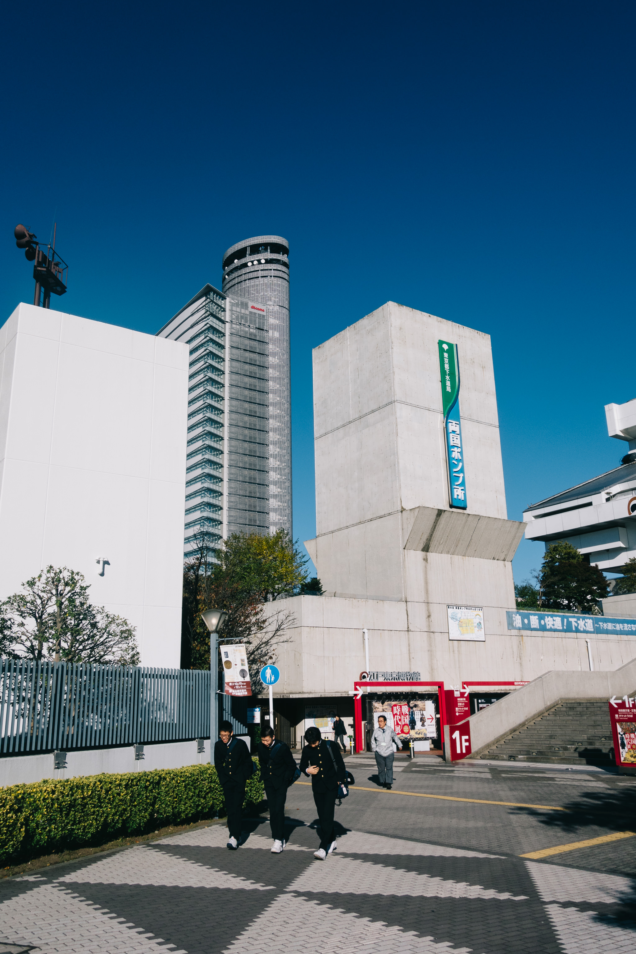 Three students in uniform walk past a modern building in Tokyo, Japan, under a clear blue sky.