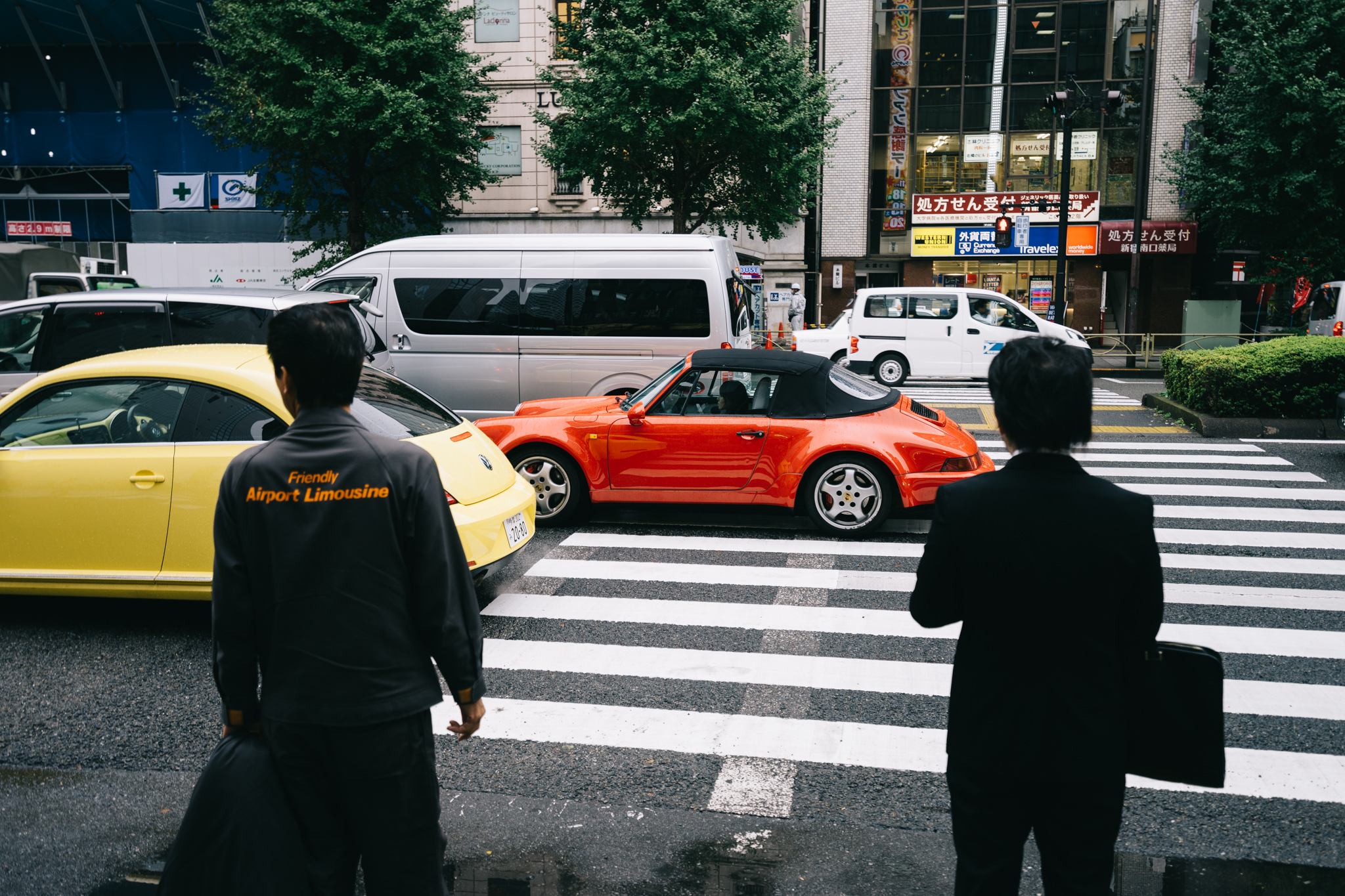 A man in a 'Friendly Airport Limousine' uniform and a businessman cross a pedestrian crossing in Japan, with a red Porsche and yellow Volkswagen in the background.