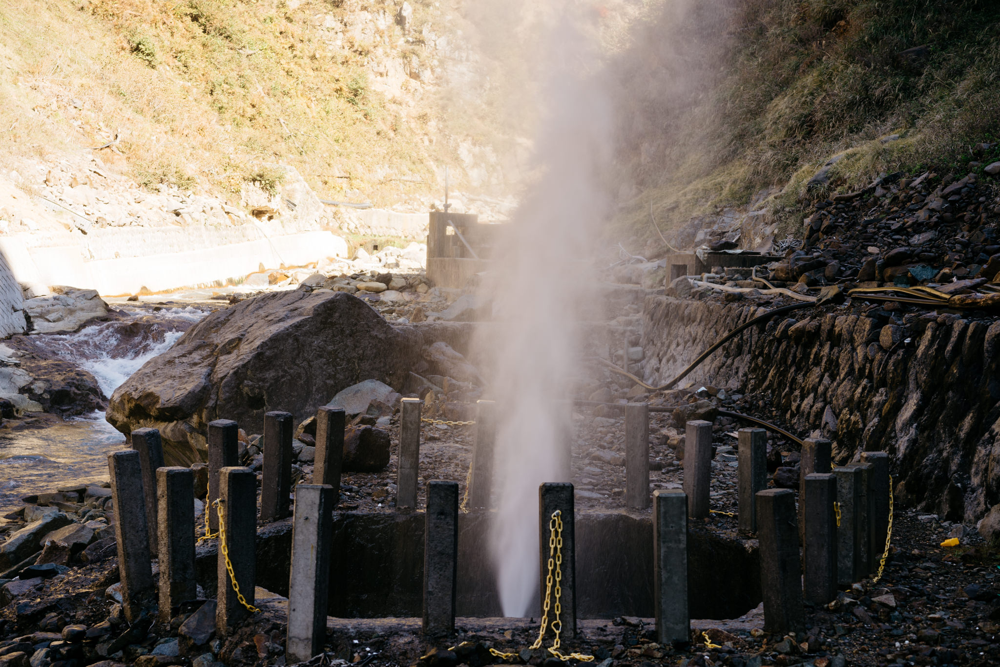 Water geyser erupting from a man-made structure near a rocky river.