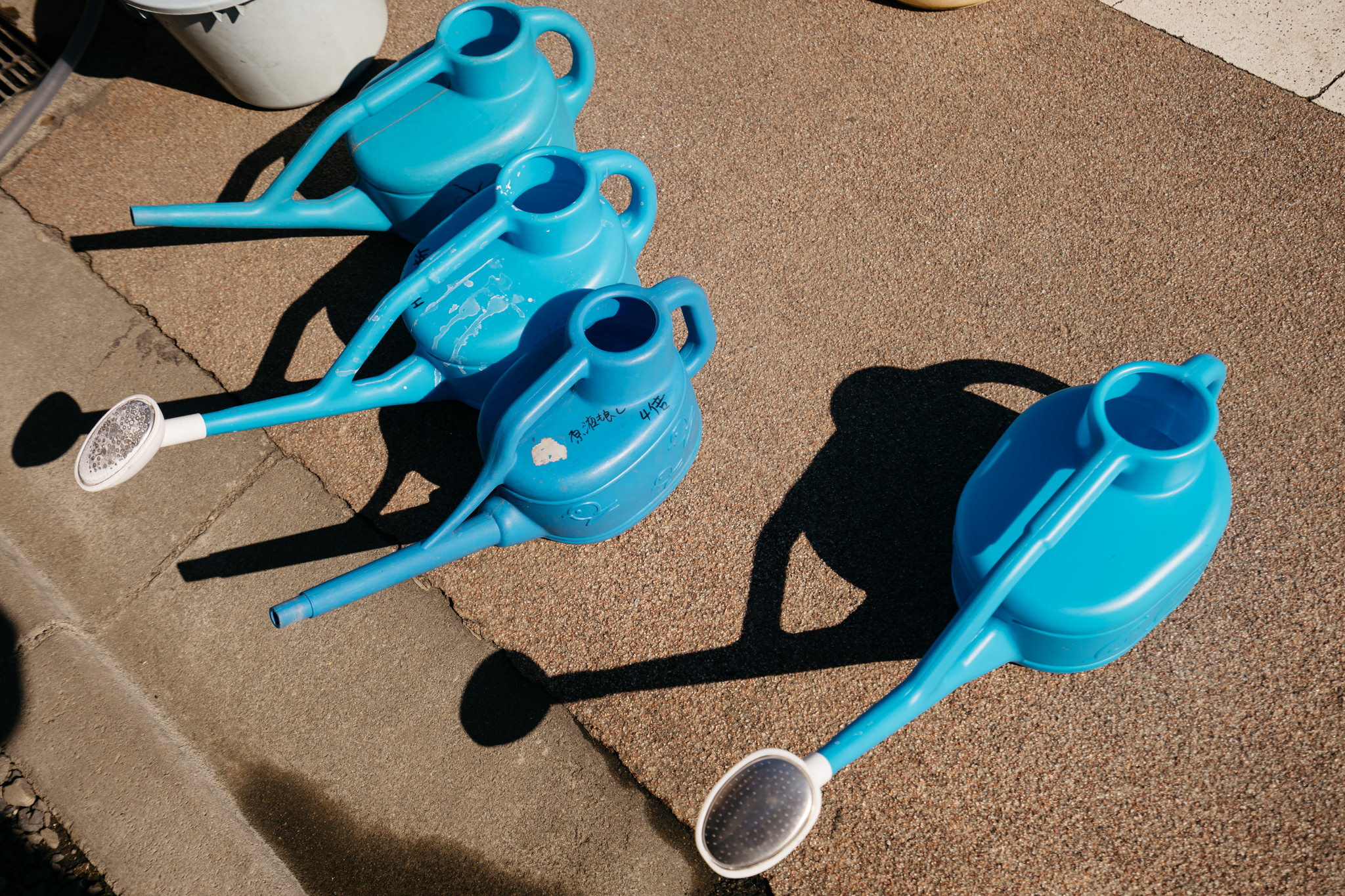 Four blue plastic watering cans on a gravel surface.