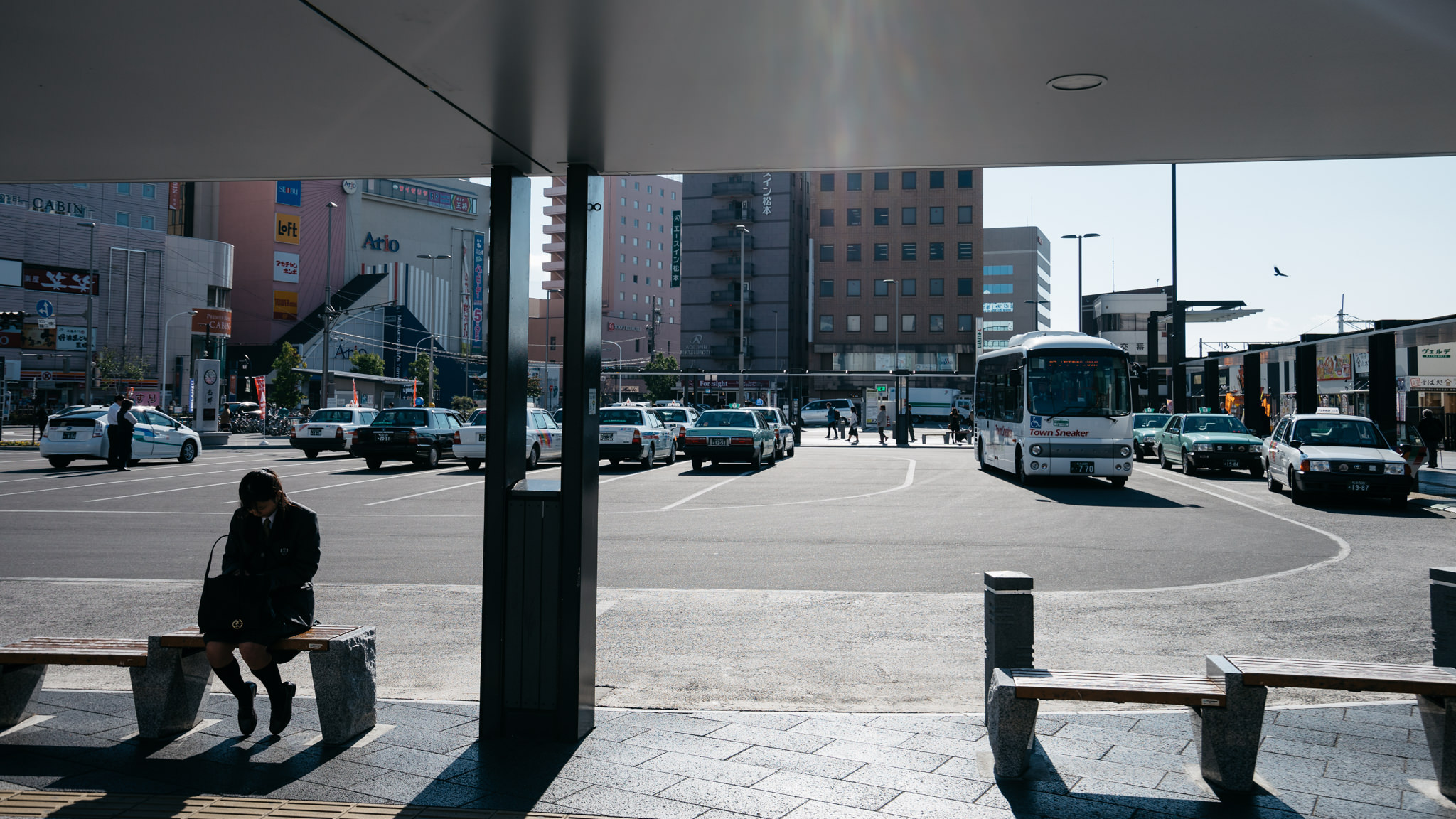 A schoolgirl sits on a bench in a Japanese city square, with taxis and a bus visible in the background.