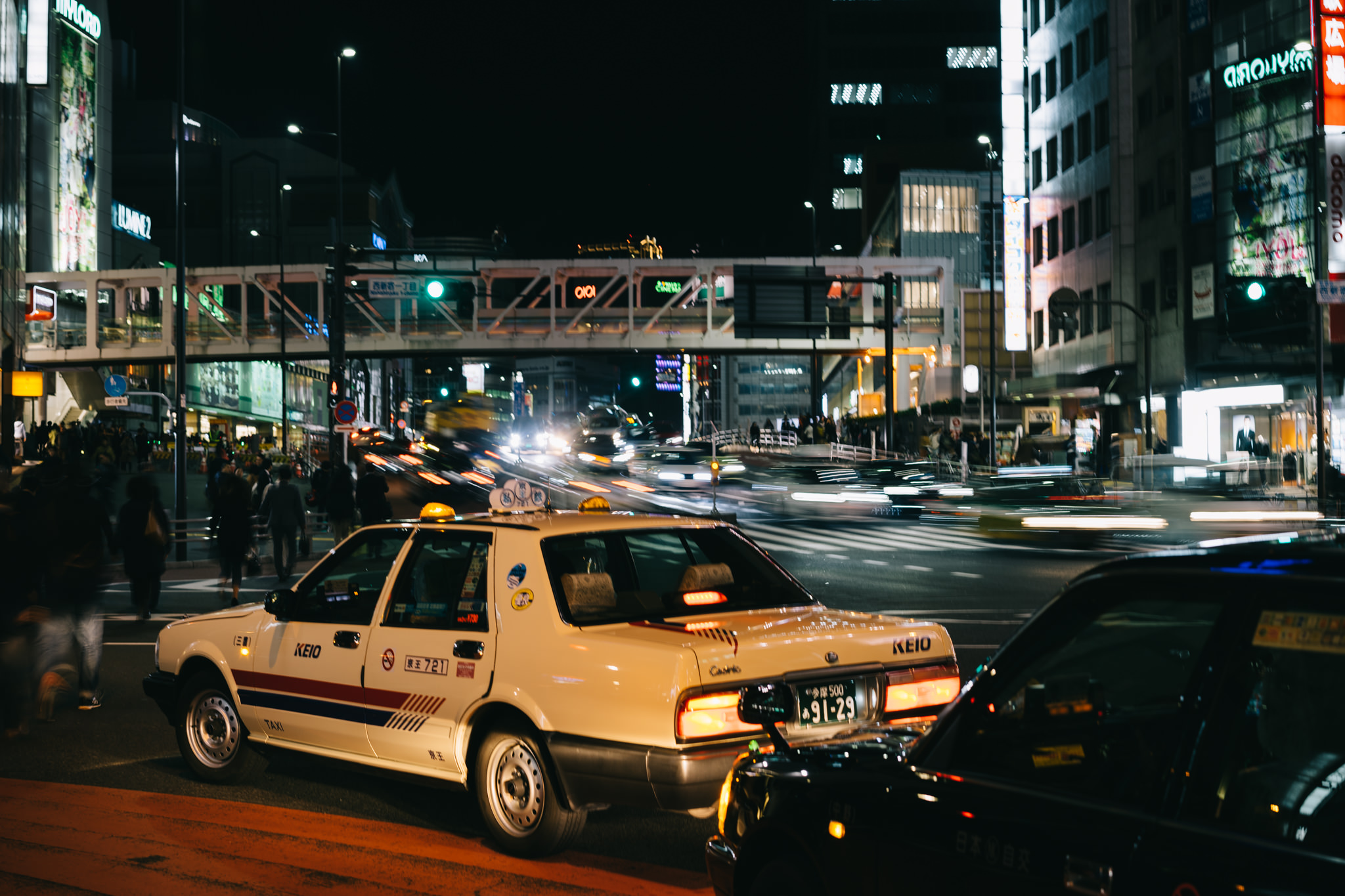 A cream-colored Keio taxi in Tokyo at night, with blurred traffic and city lights.