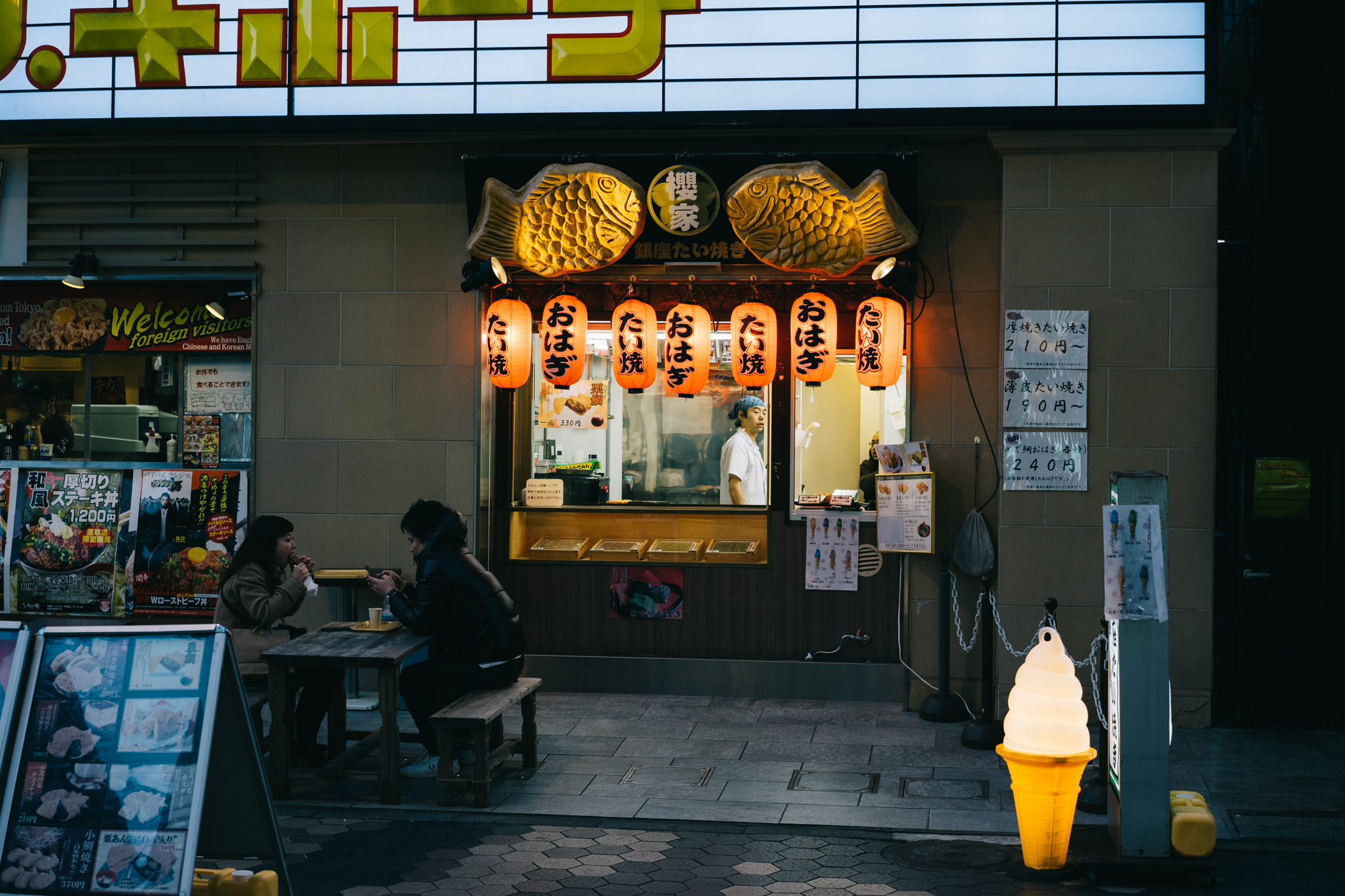Tokyo street scene at night; two people eating outside a taiyaki restaurant.