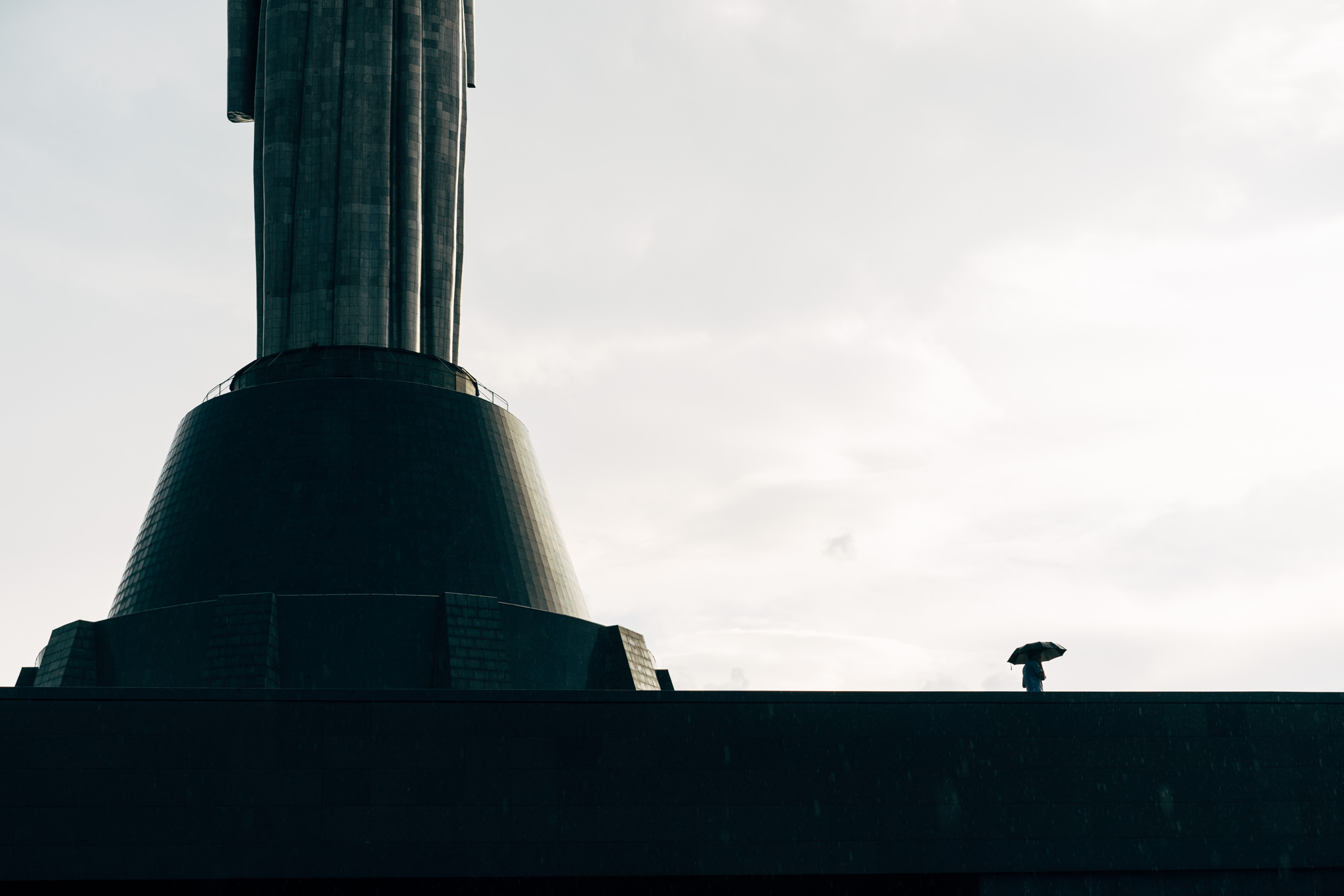 Motherland Monument in Kyiv, Ukraine, with a person holding an umbrella in the distance.