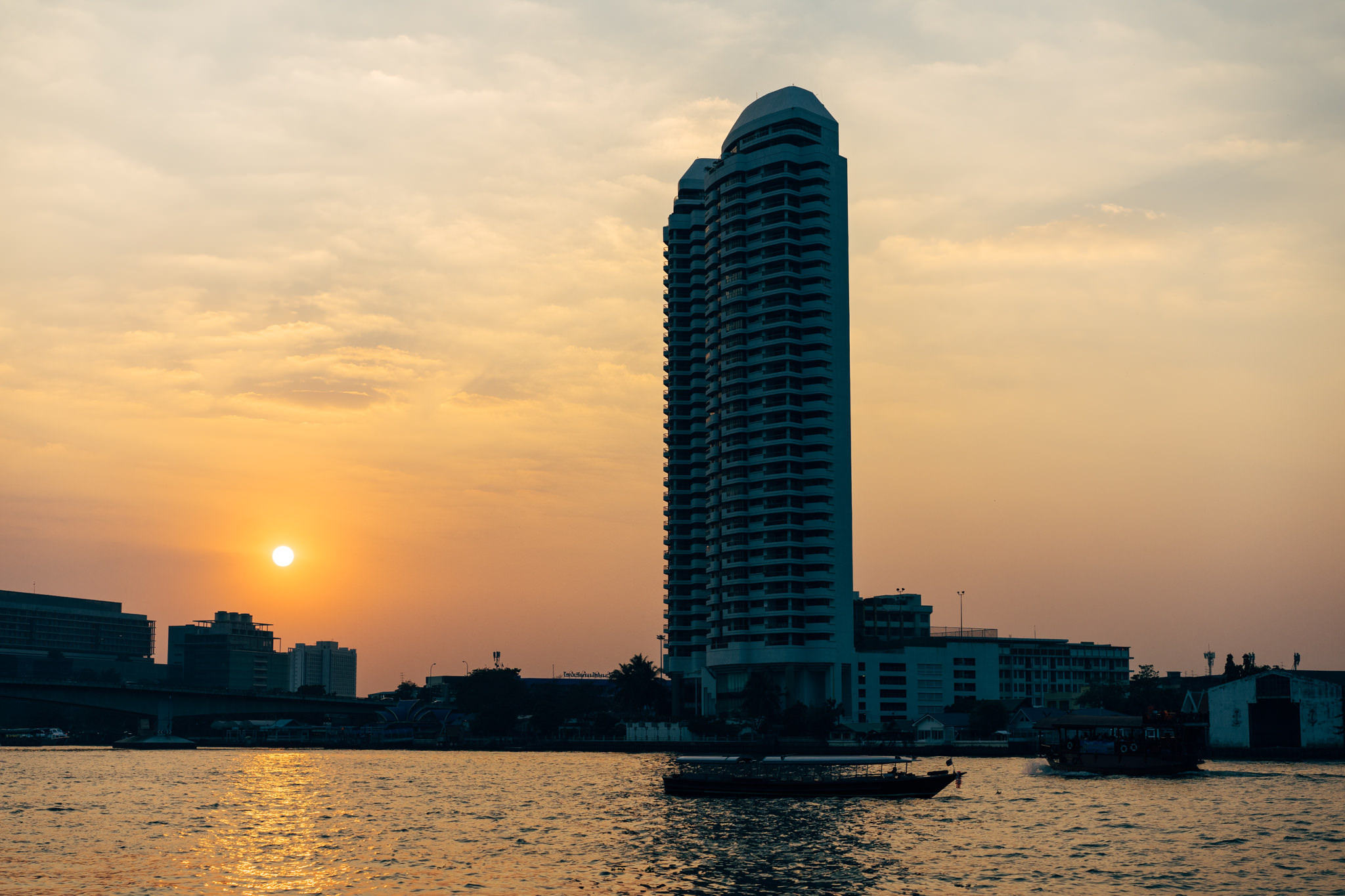 Bangkok sunset, tall building silhouetted against orange sky, river with boats.