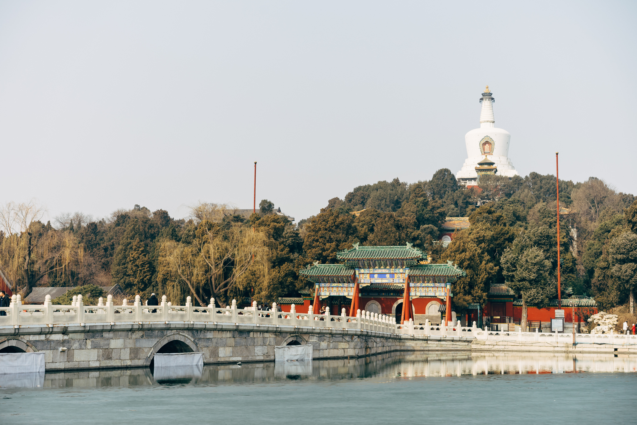Beihai Park in Beijing, featuring a stone bridge over a partially frozen lake and a white pagoda on a hill in the background.