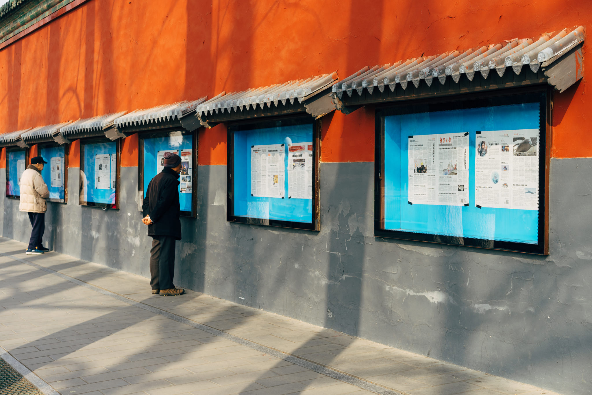 Two people reading newspapers posted on a wall in Beihai Park, Beijing.